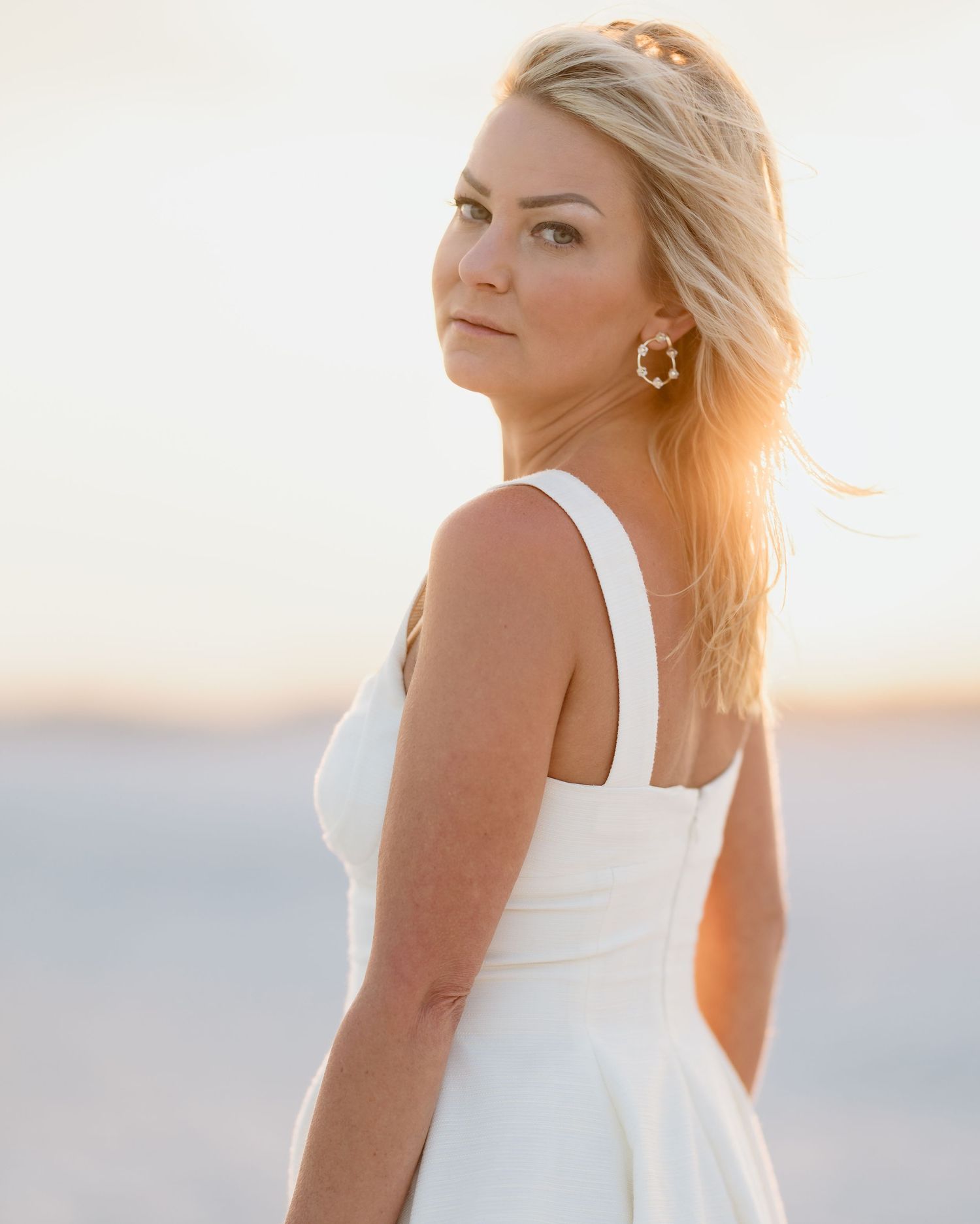 Woman in white dress with gold earrings at sunset beach, blonde hair blowing in breeze, golden hour portrait.