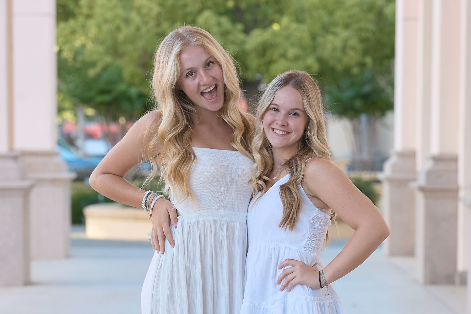 Four people in white sundresses pose together outdoors against a blurred architectural backdrop.