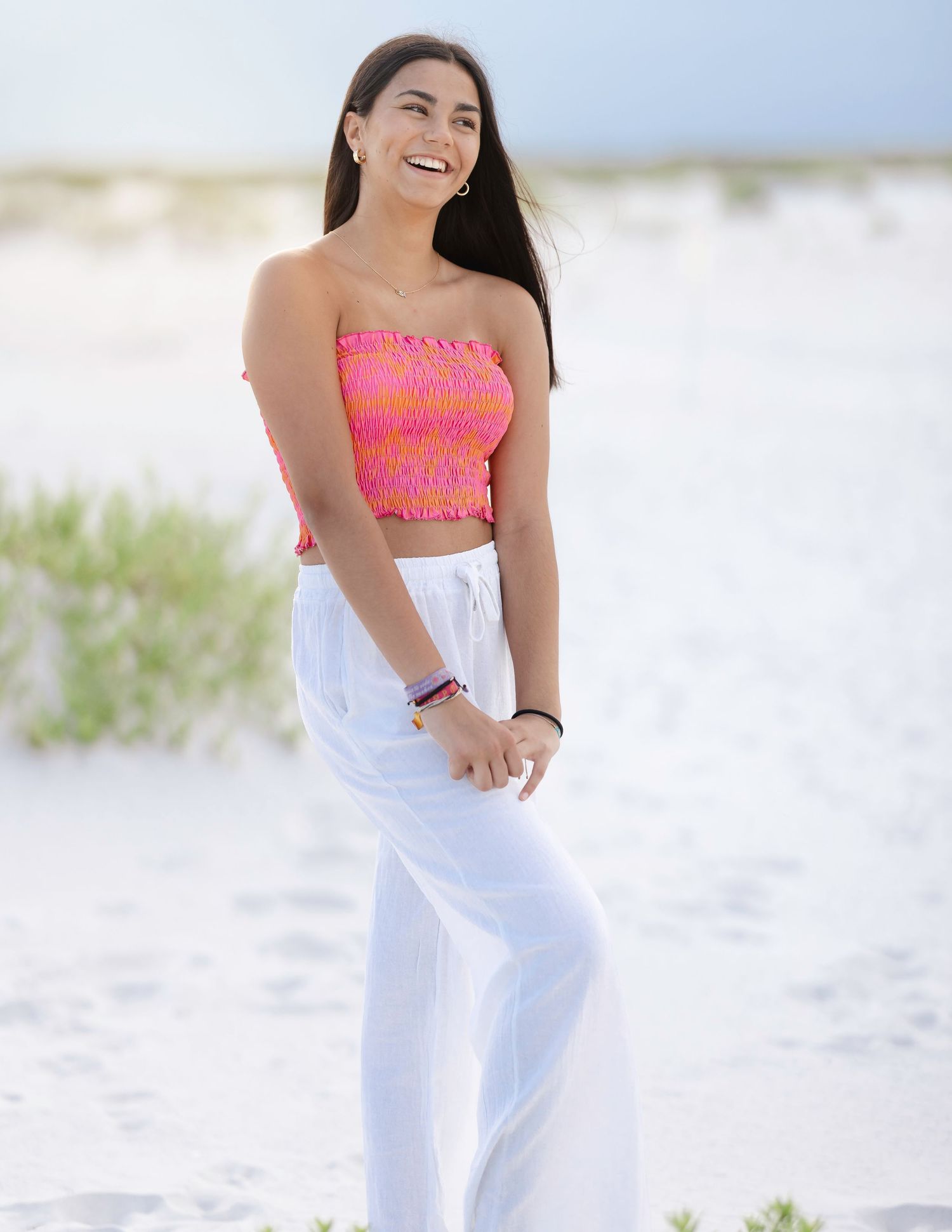 Woman in vibrant coral top poses energetically on sunlit beach.