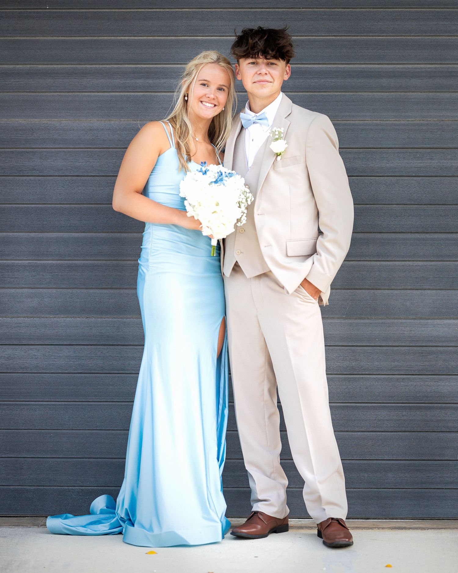 A couple in formal attire poses together against a gray wall, with the woman in a flowing light blue gown.