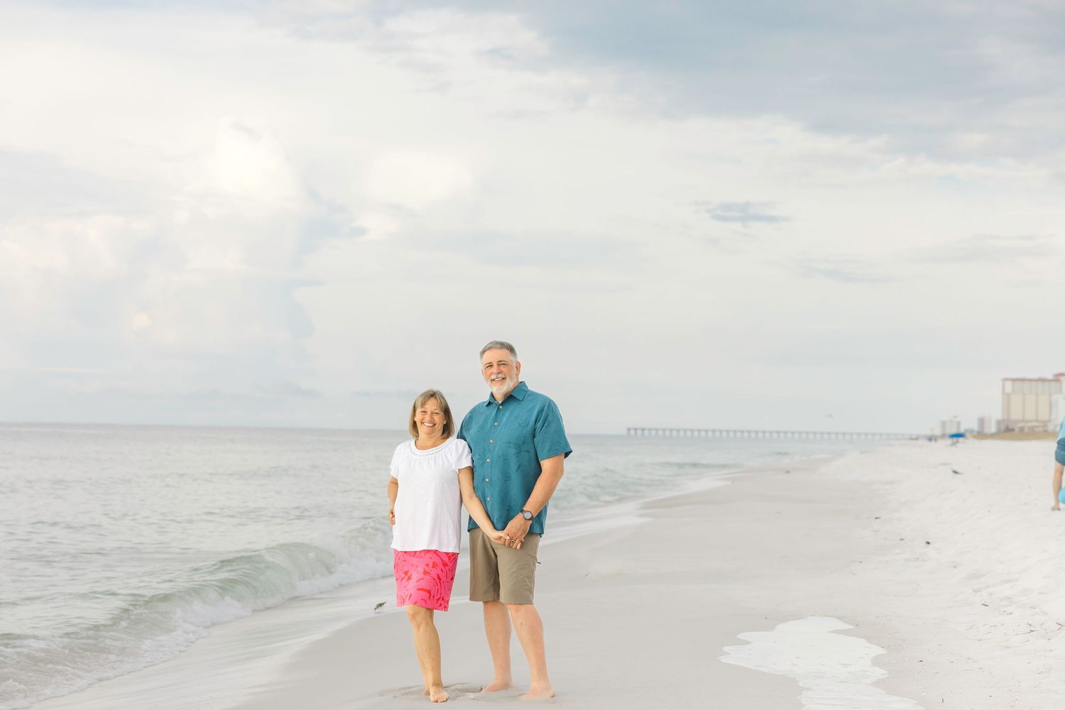 Two people walk along a white sandy beach at sunset, holding hands and strolling near the ocean's edge.