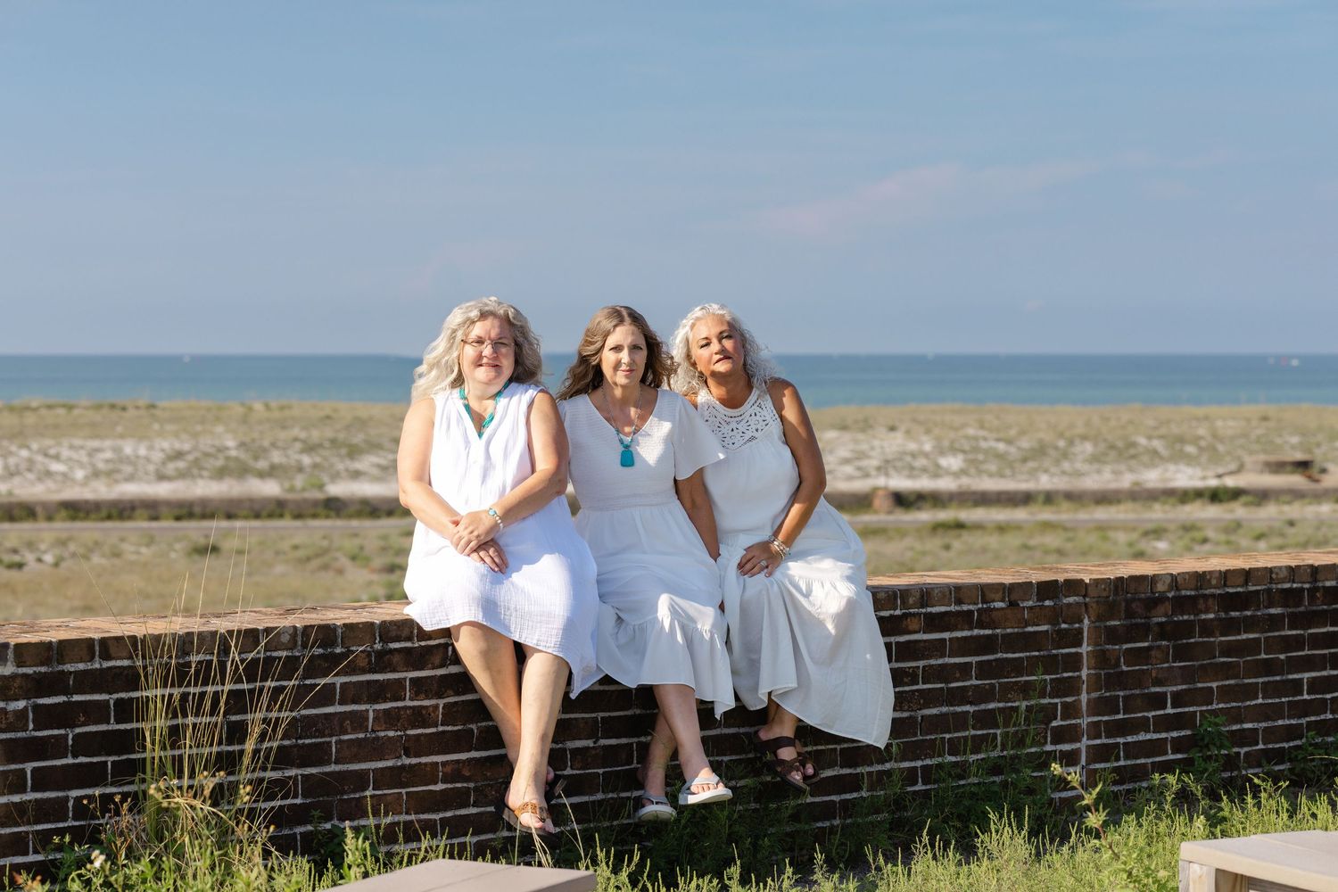Three friends in white dresses sit together on a brick wall by the beach on a sunny summer day.