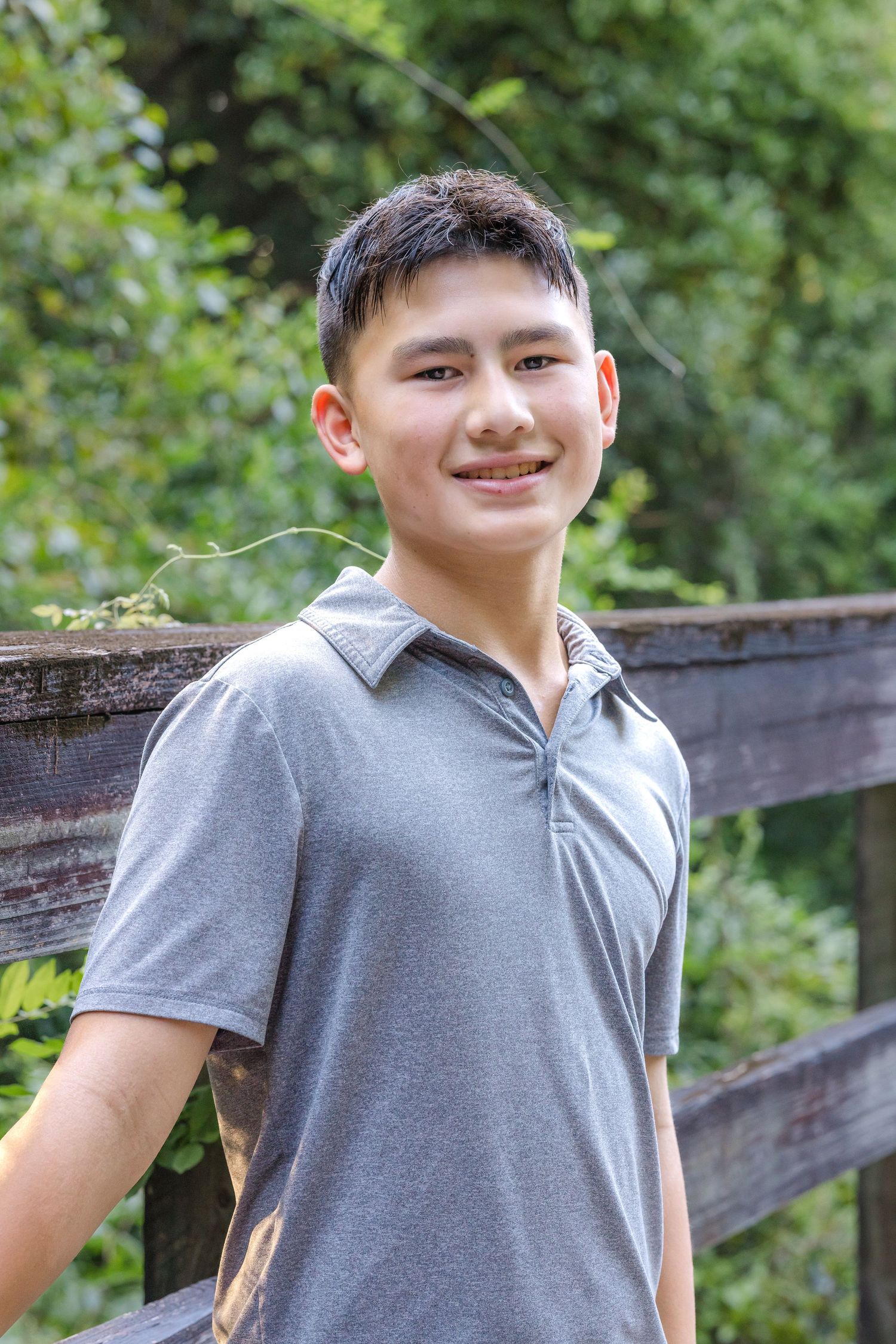 Young person in grey polo shirt smiling outdoors by wooden fence with green foliage in background.