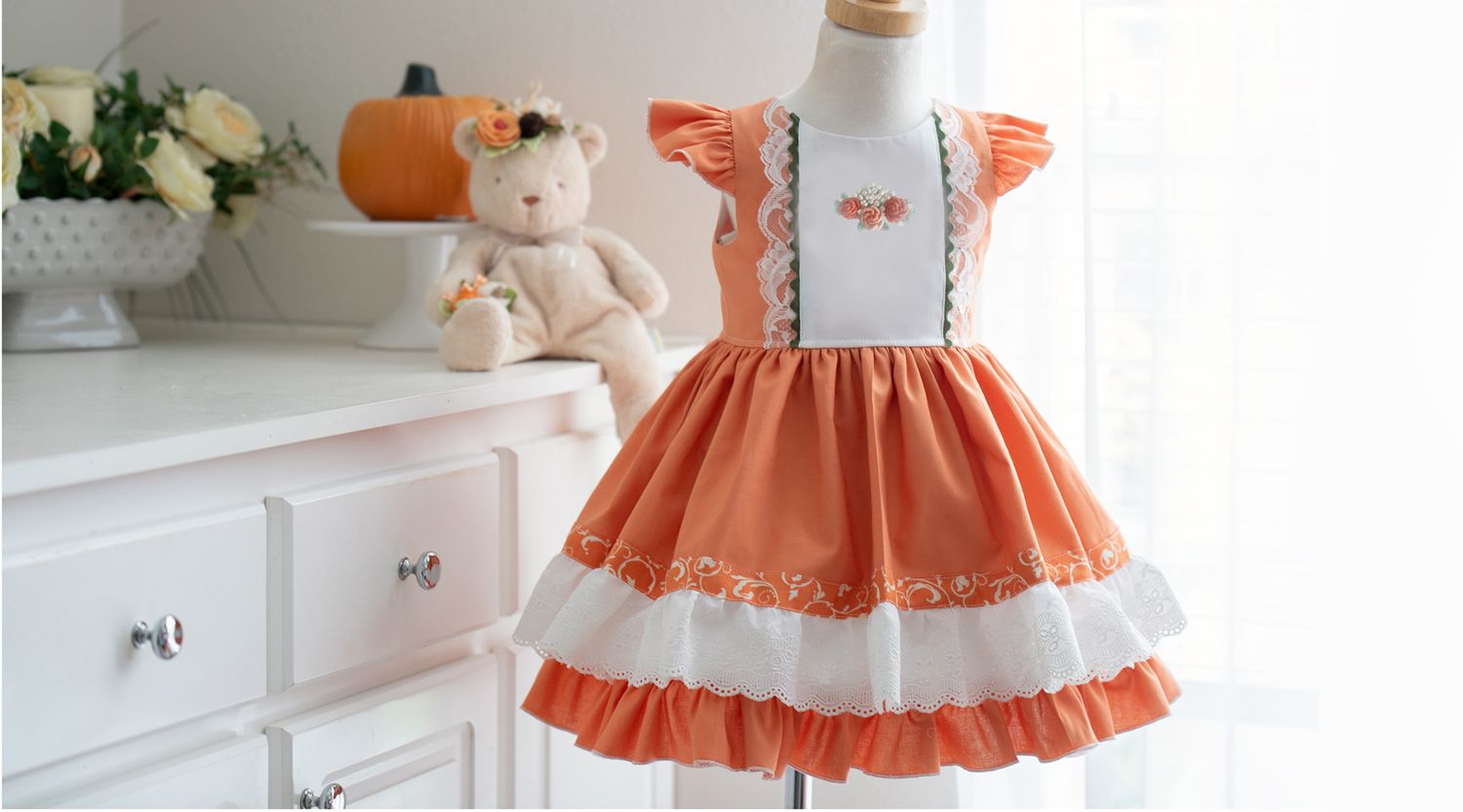 Orange and white vintage-style children's dress with ruffles and suspenders hangs beside a plush teddy bear on dresser.