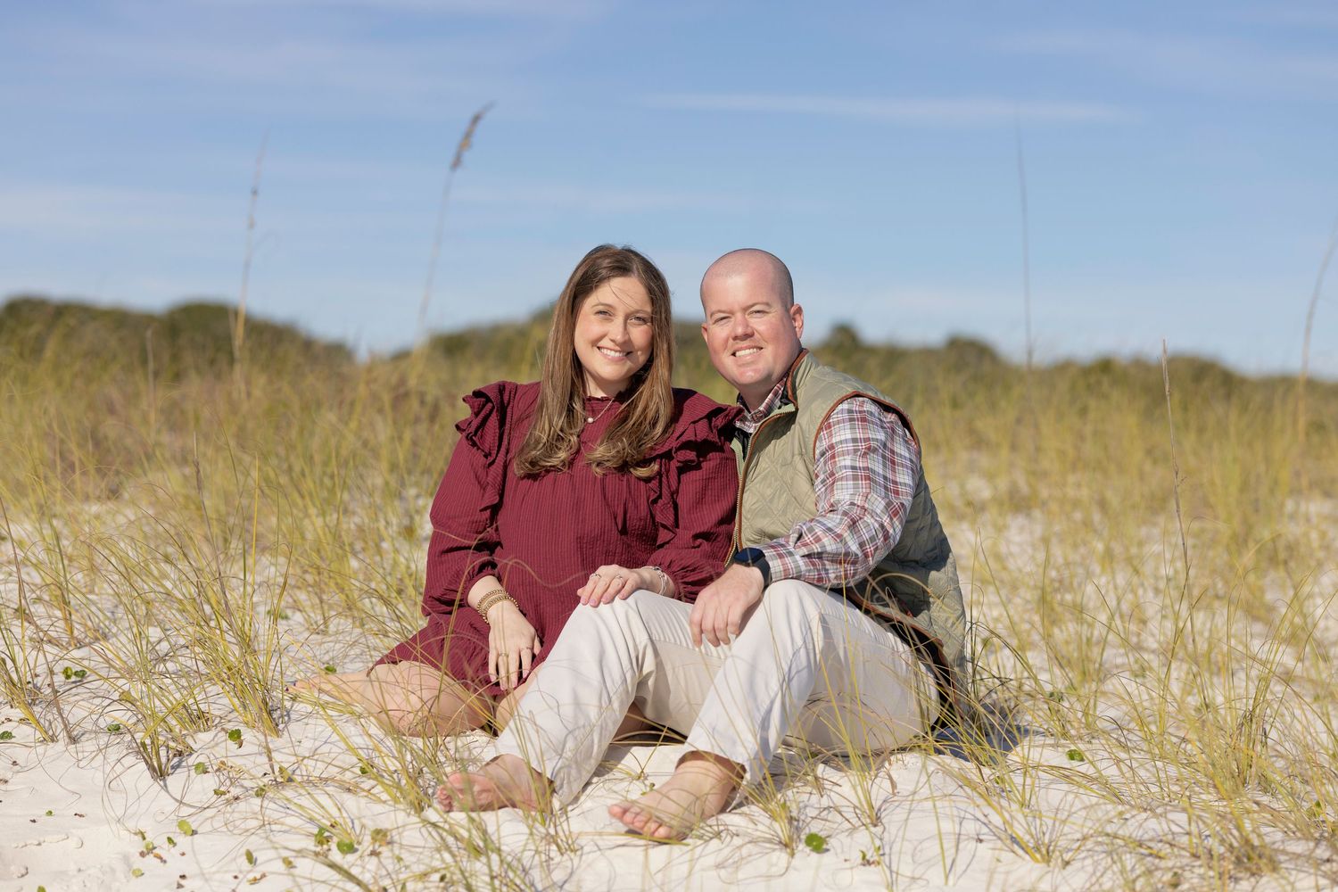 Couple sitting together on white sand beach dunes with sea oats, smiling outdoors under blue sky.