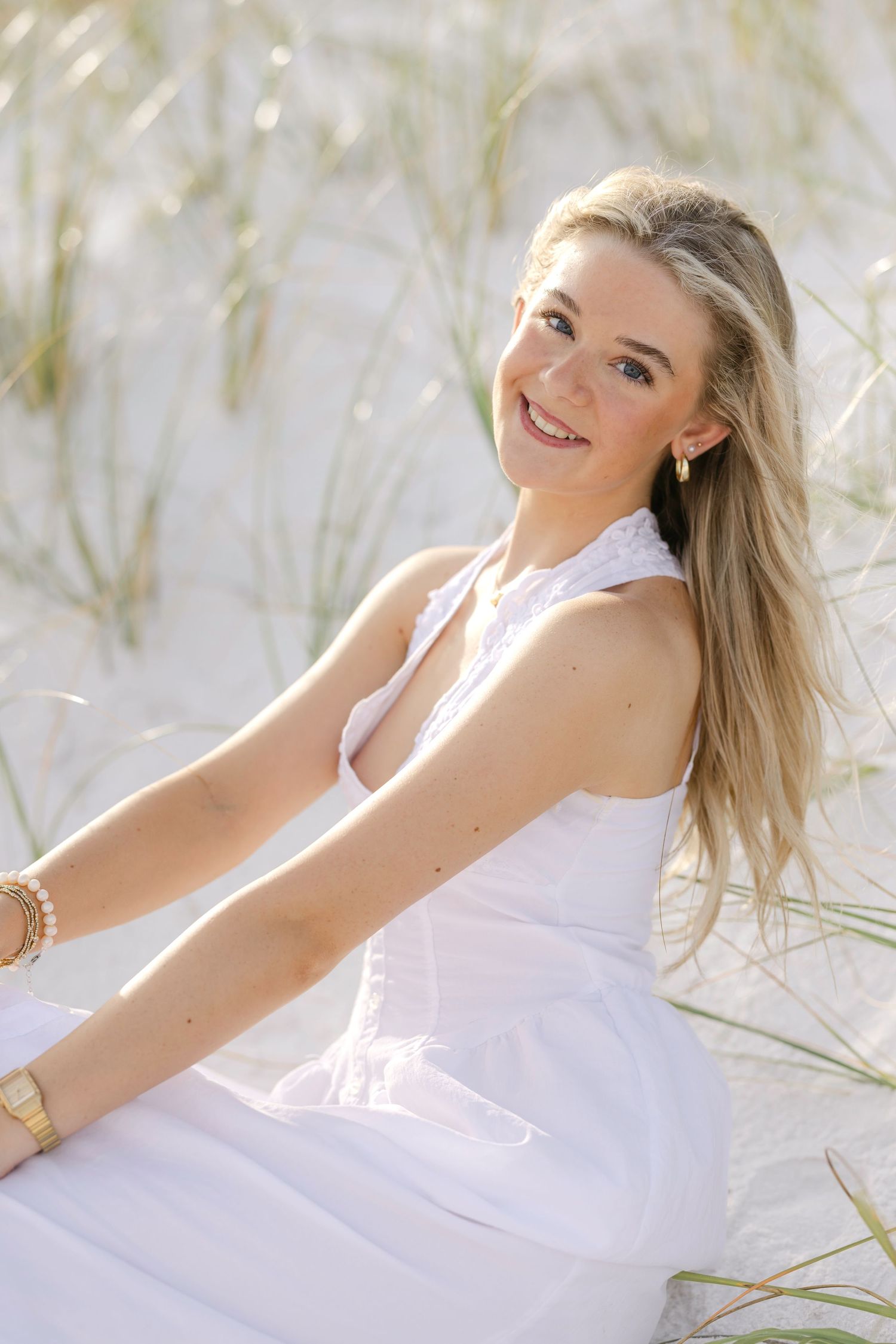 Smiling blonde woman in white halter dress sitting on sandy beach dunes with tall grass in background.