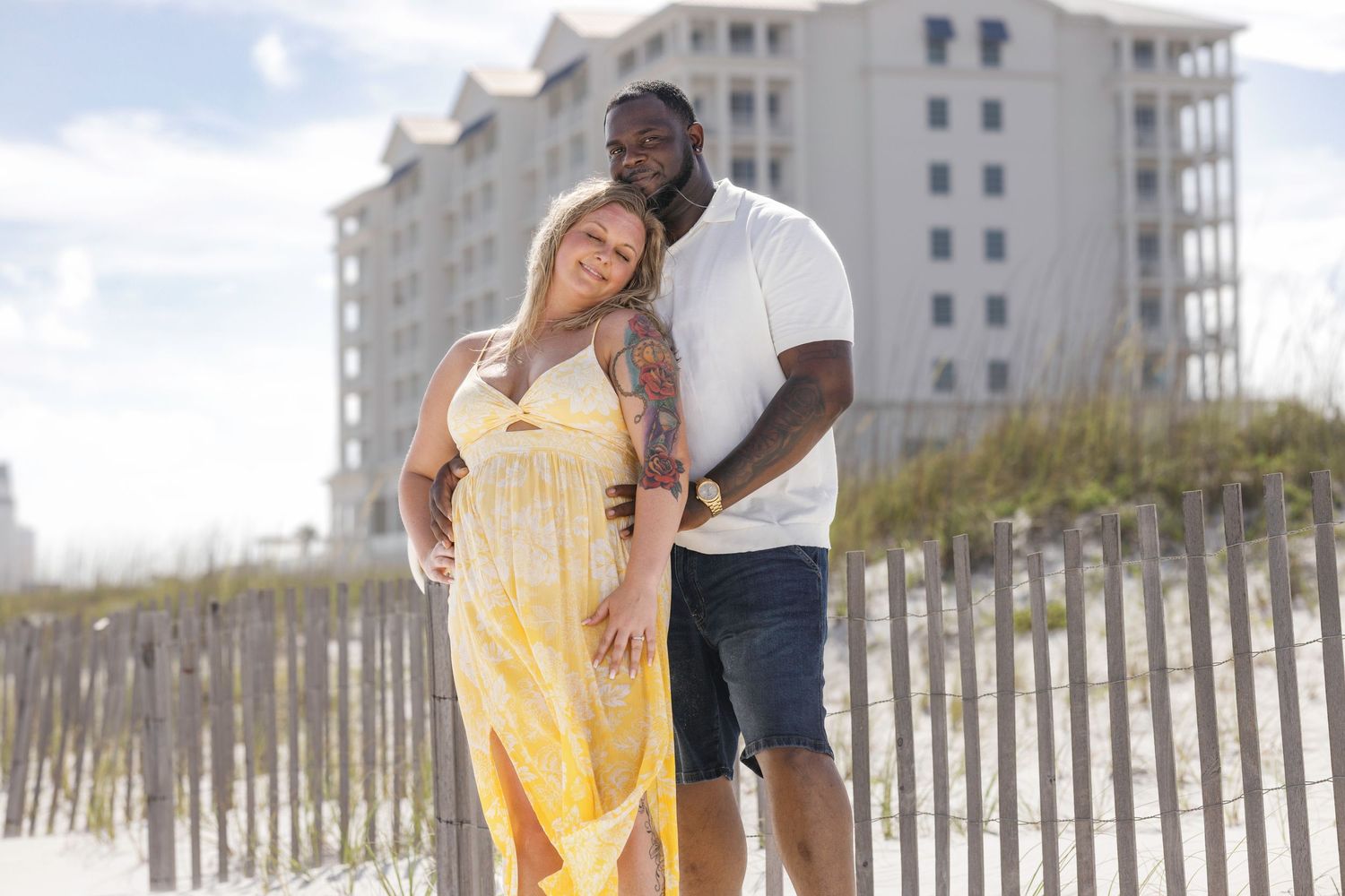 Couple embracing on beach boardwalk, woman in yellow floral dress, man in white shirt, hotel in background.