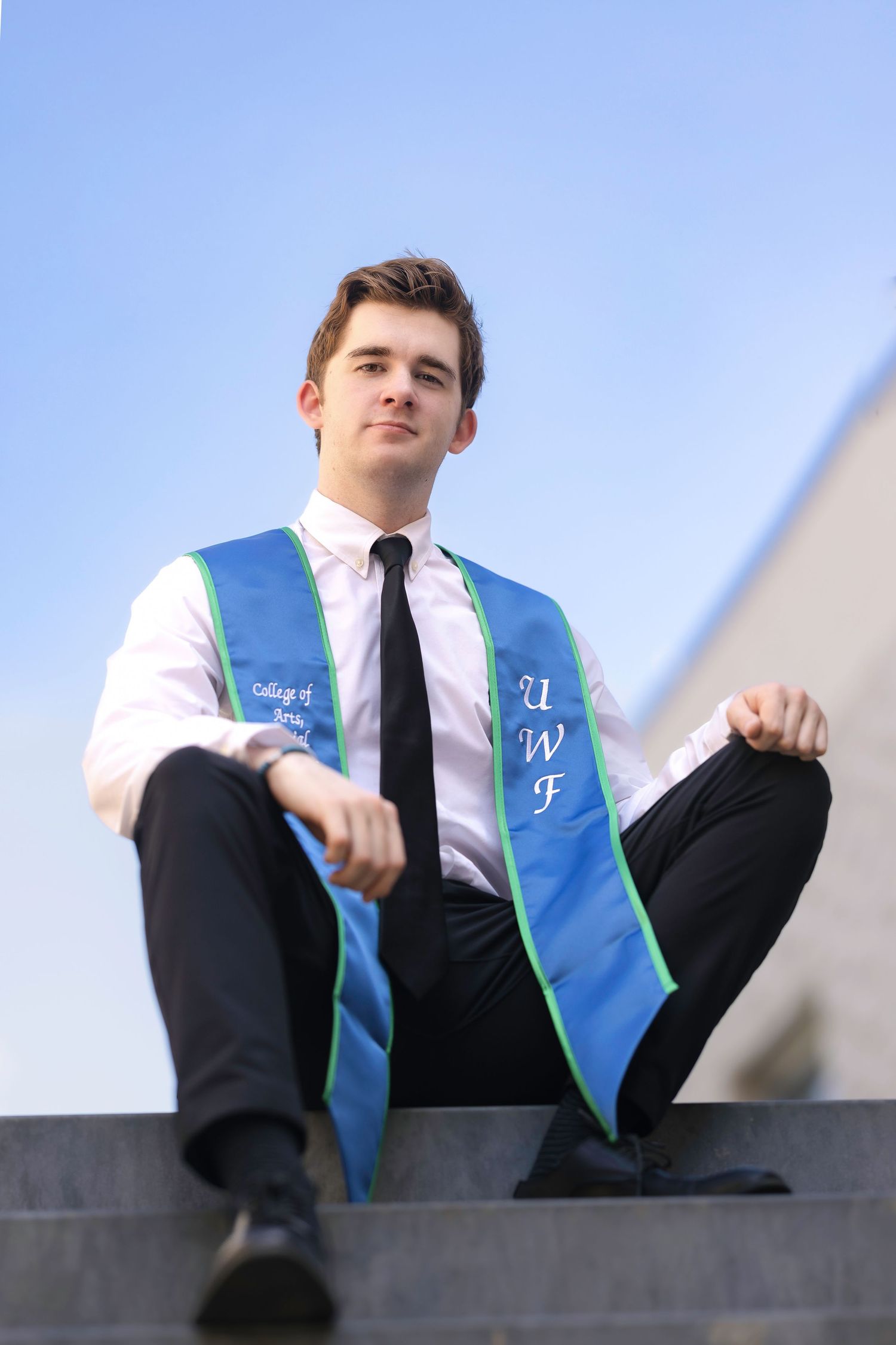 Young man in white shirt, black tie, and UWF graduation sash sits on steps outdoors under blue sky.