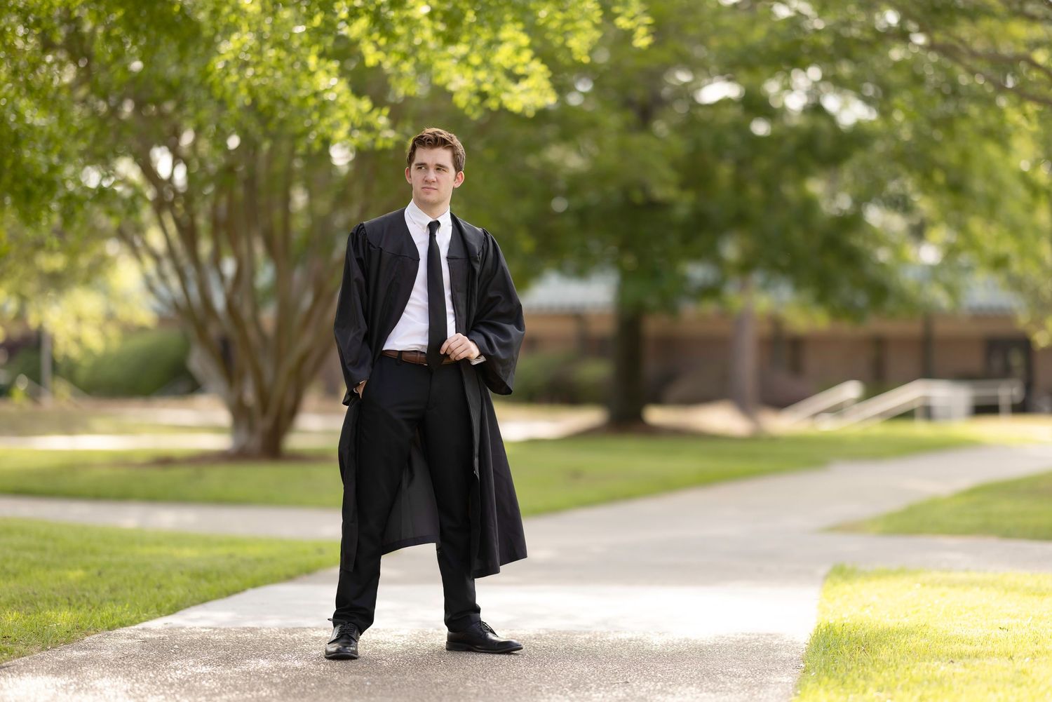Young man in graduation gown and suit standing confidently on a campus pathway surrounded by green trees.
