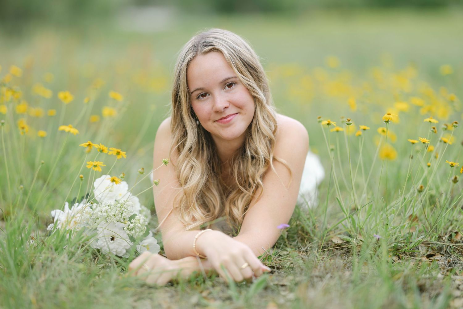 A young blonde woman lies in a wildflower meadow holding a white bouquet on a sunny day.