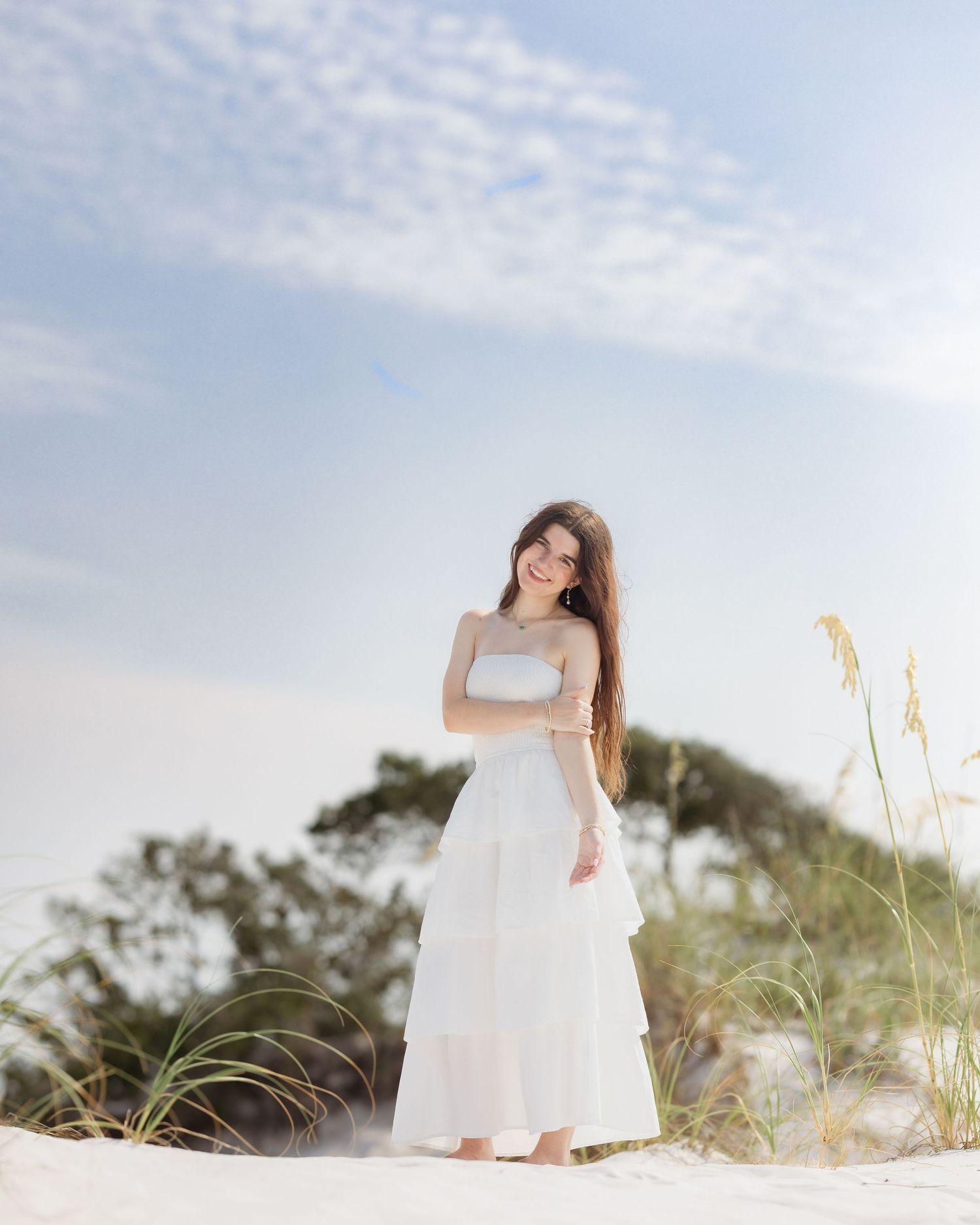 Smiling young woman in white tiered dress standing on white sand beach under blue sky.