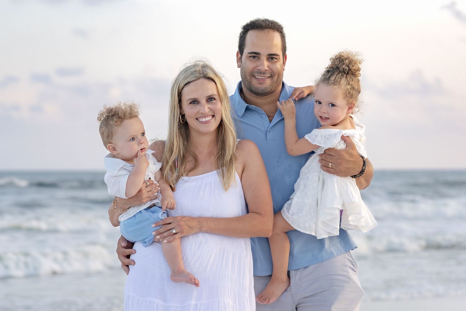 Family of four posing on beach, parents holding toddlers, smiling, ocean waves in background.