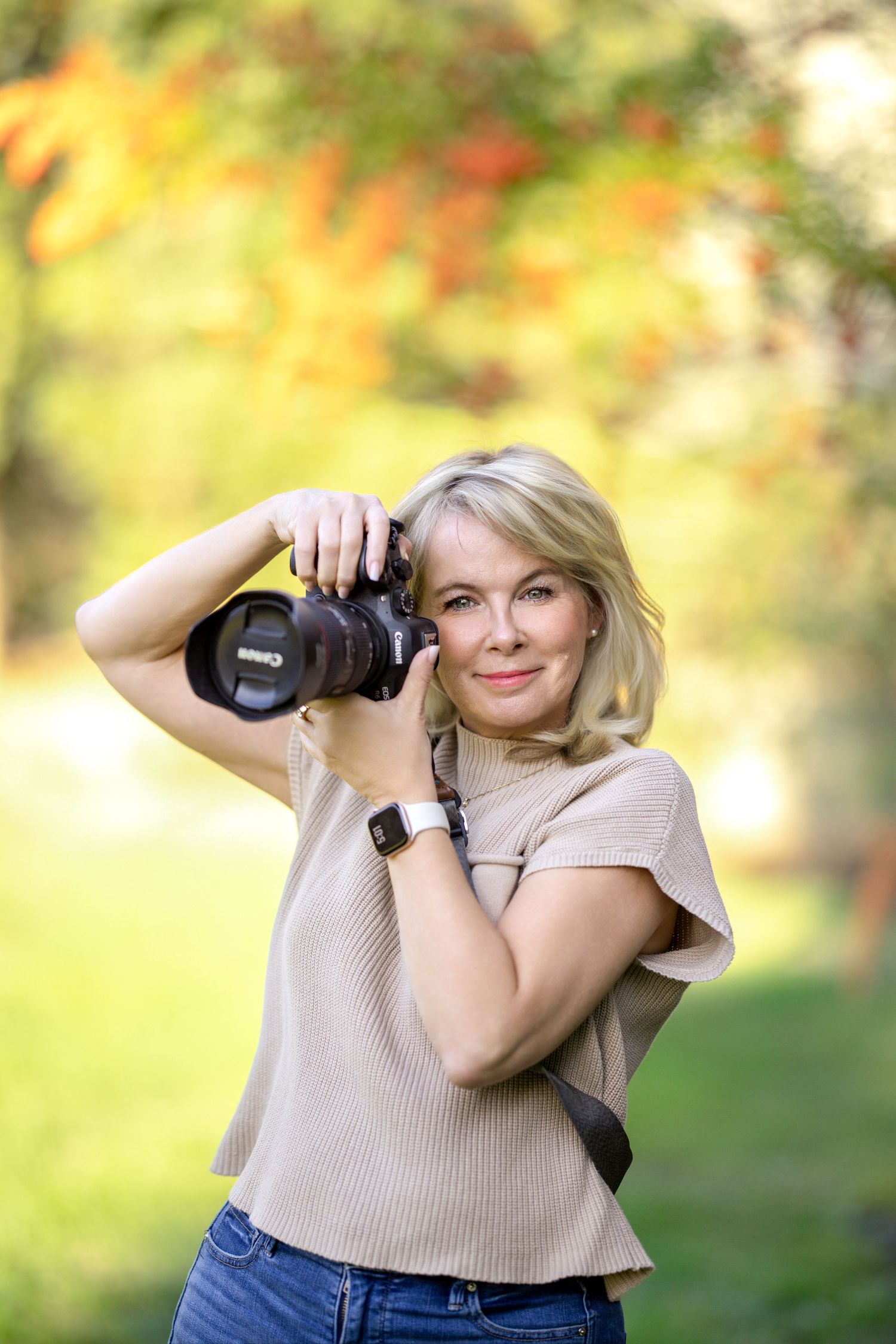 A photographer holds up a professional camera against an autumn background with soft bokeh lighting effects.