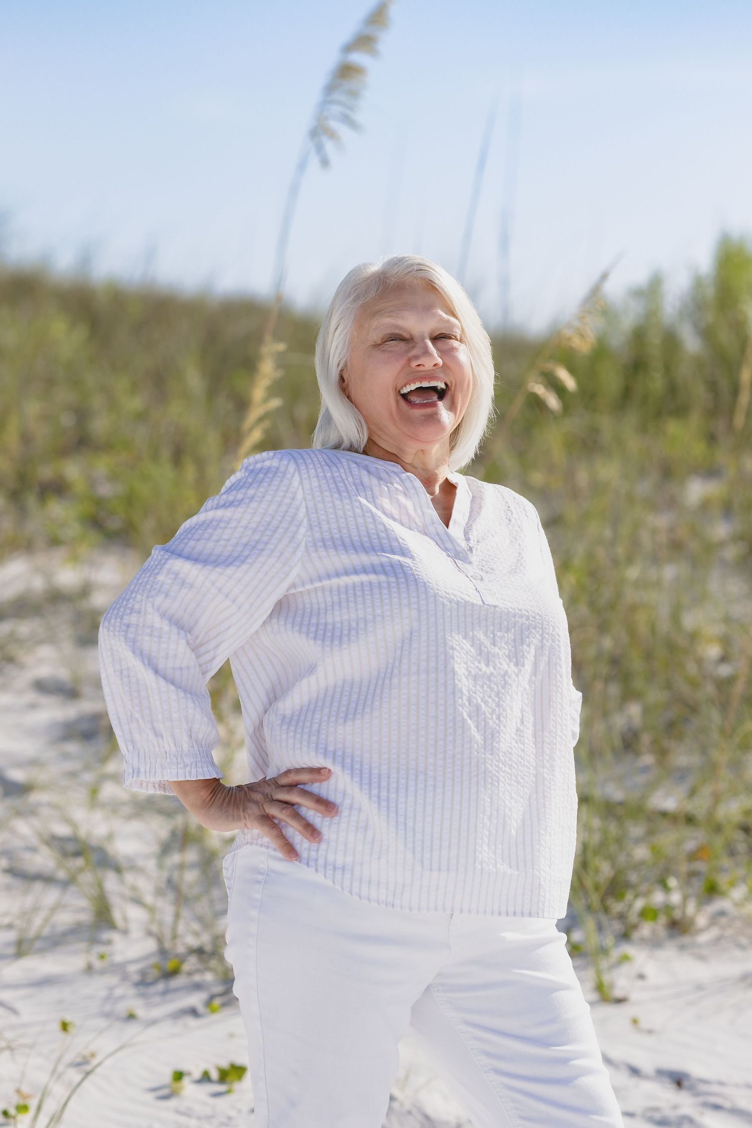Cheerful senior woman with white hair laughing on sandy beach dunes, wearing white outfit on sunny day.