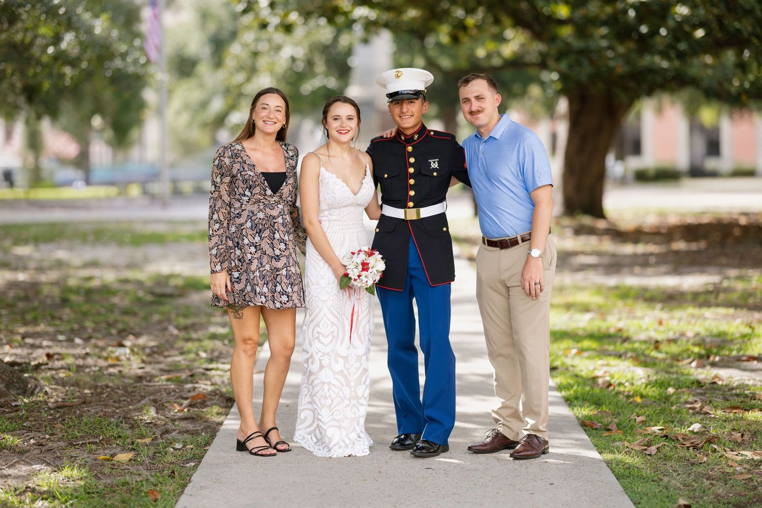 A bride in a white dress and groom in Marine dress blues pose with two guests outdoors on a sunny day.