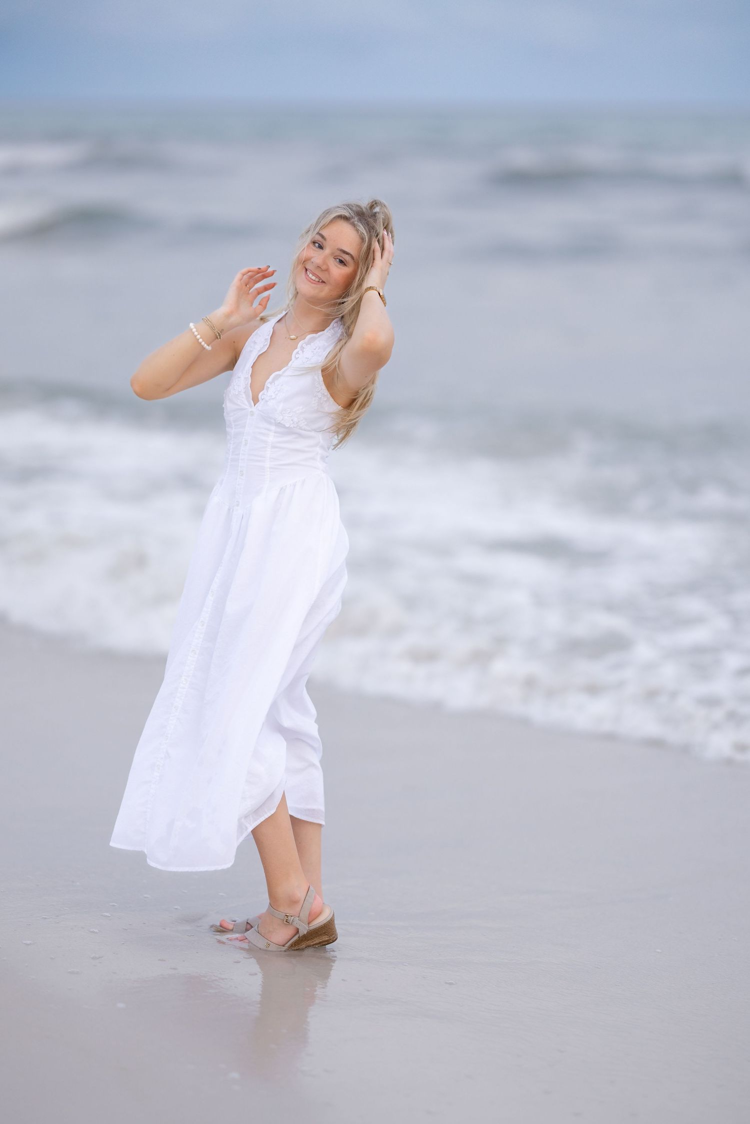 Joyful blonde woman in white dress dancing on ocean shoreline waves.