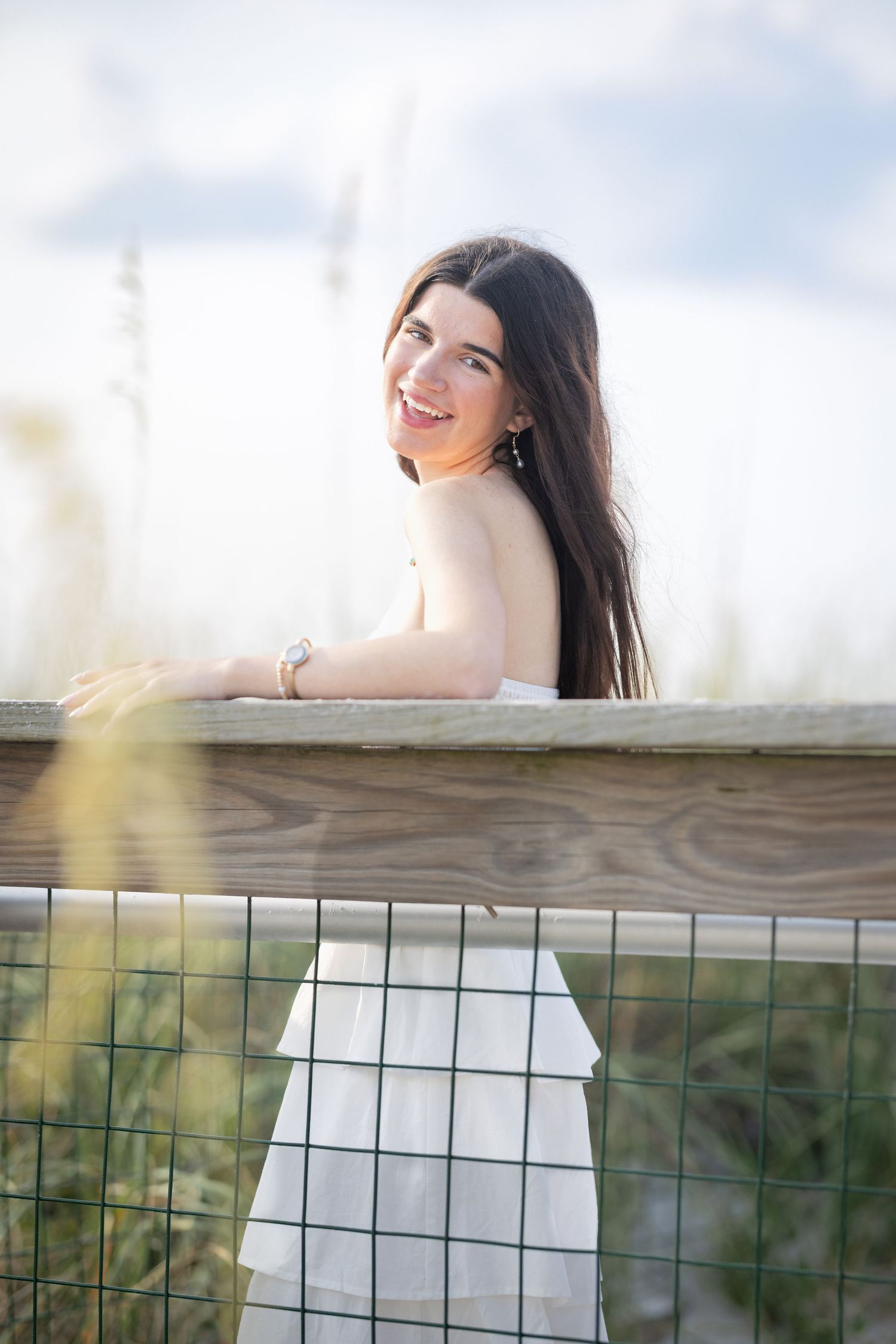 Smiling brunette woman in white dress leaning on wooden beach fence railing outdoors.