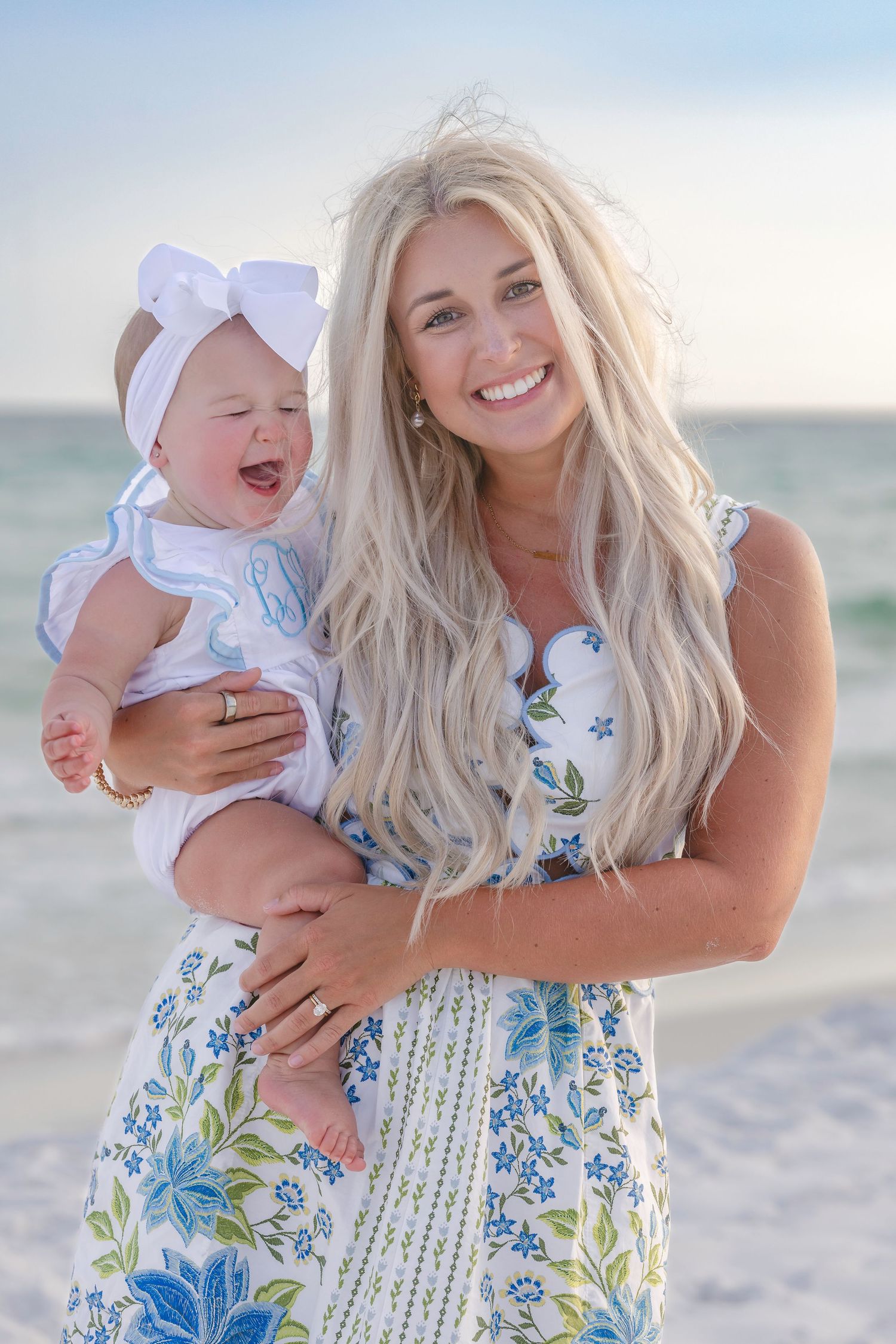 Smiling blonde mom holding laughing baby girl in white bow headband on sunny beach.