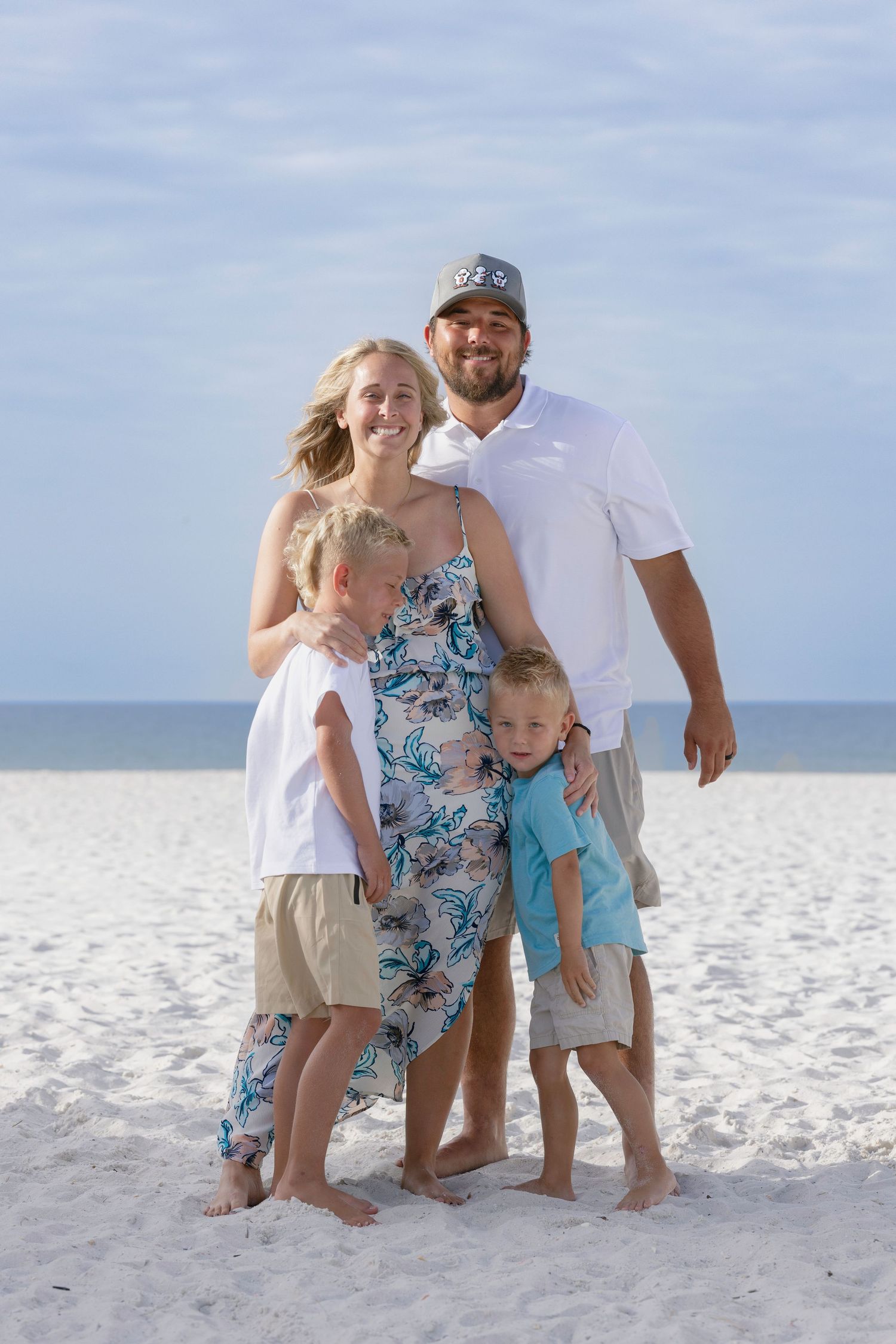 Family of four posing on white sand beach, parents smiling with two young boys hugging them.