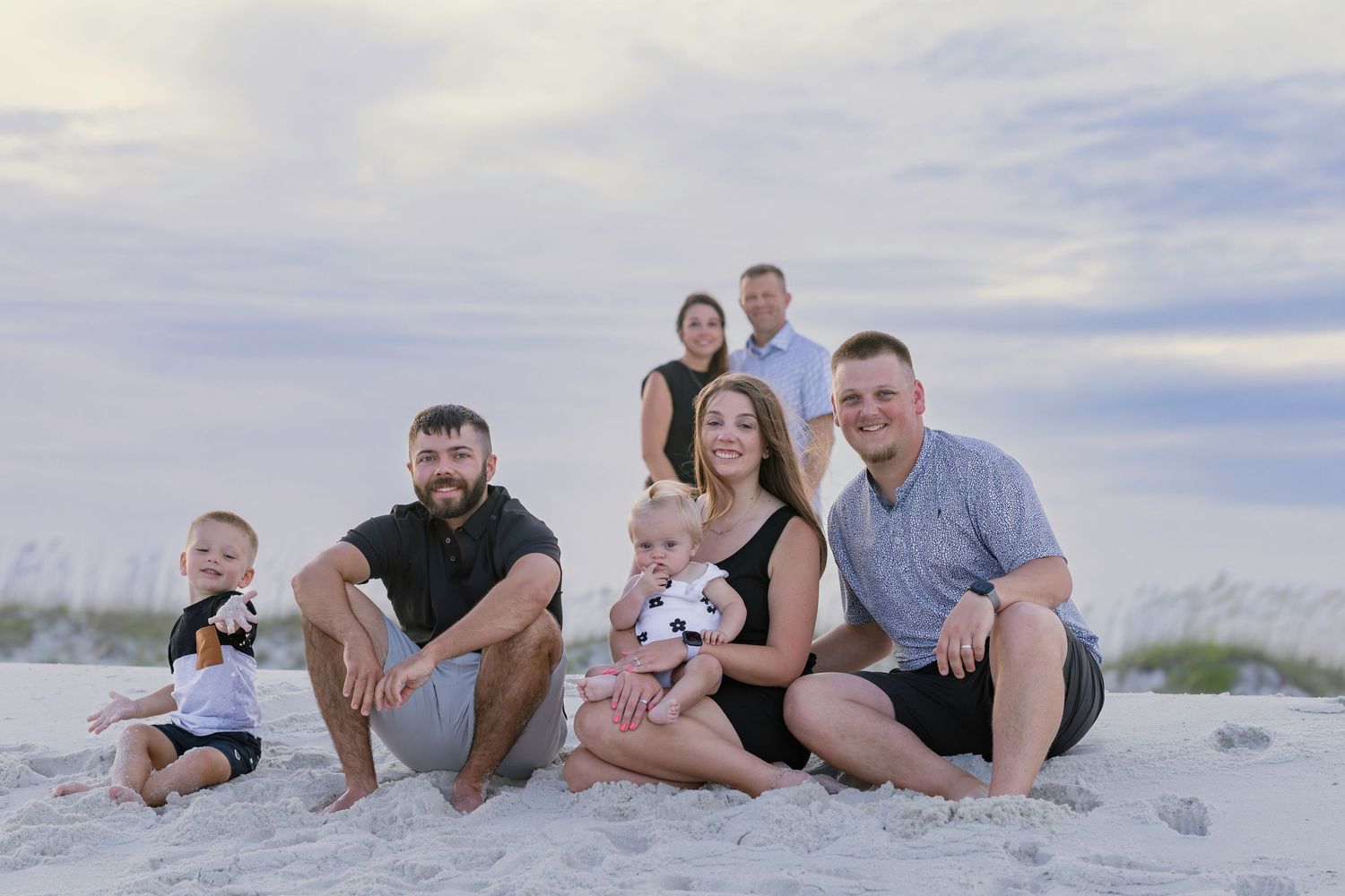 Family group portrait sitting on sandy beach with sea oats and cloudy sky backdrop.
