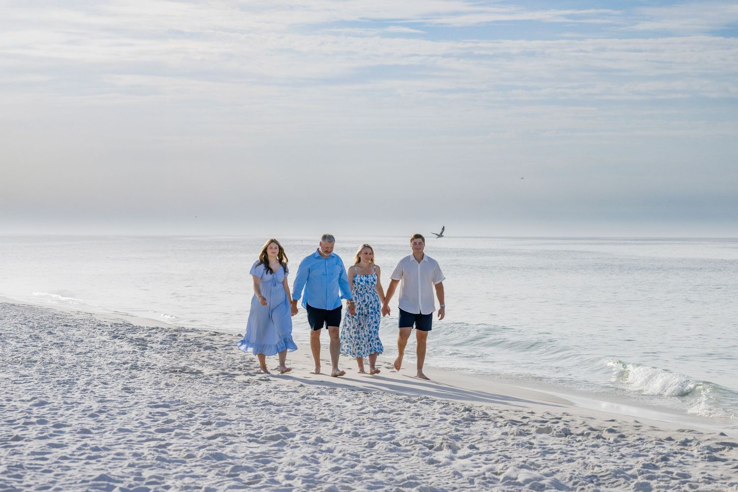 Family of four walking hand-in-hand on a serene white sand beach with calm ocean waves in the background.