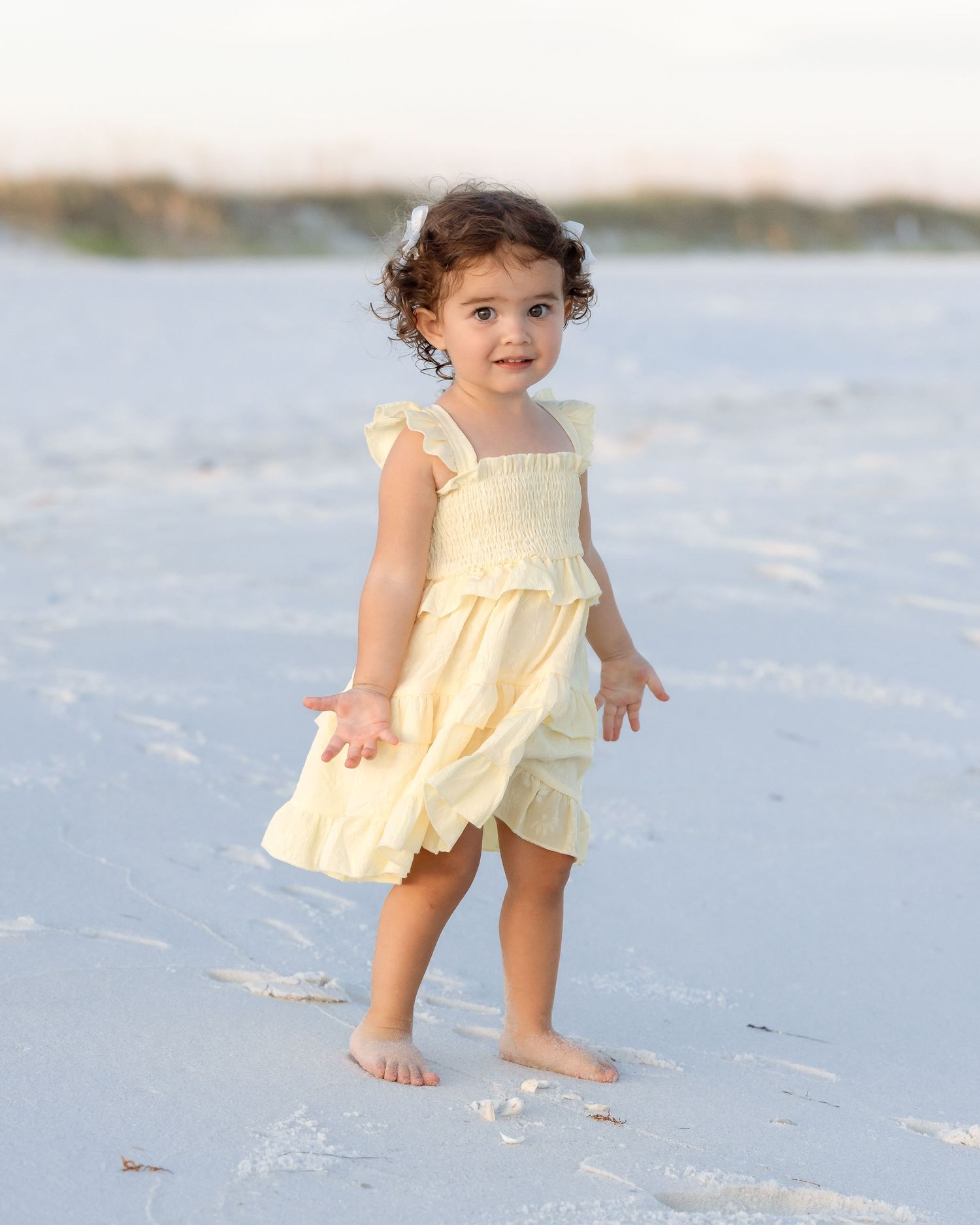 Adorable toddler girl in yellow dress walking barefoot on white sand beach at sunset.