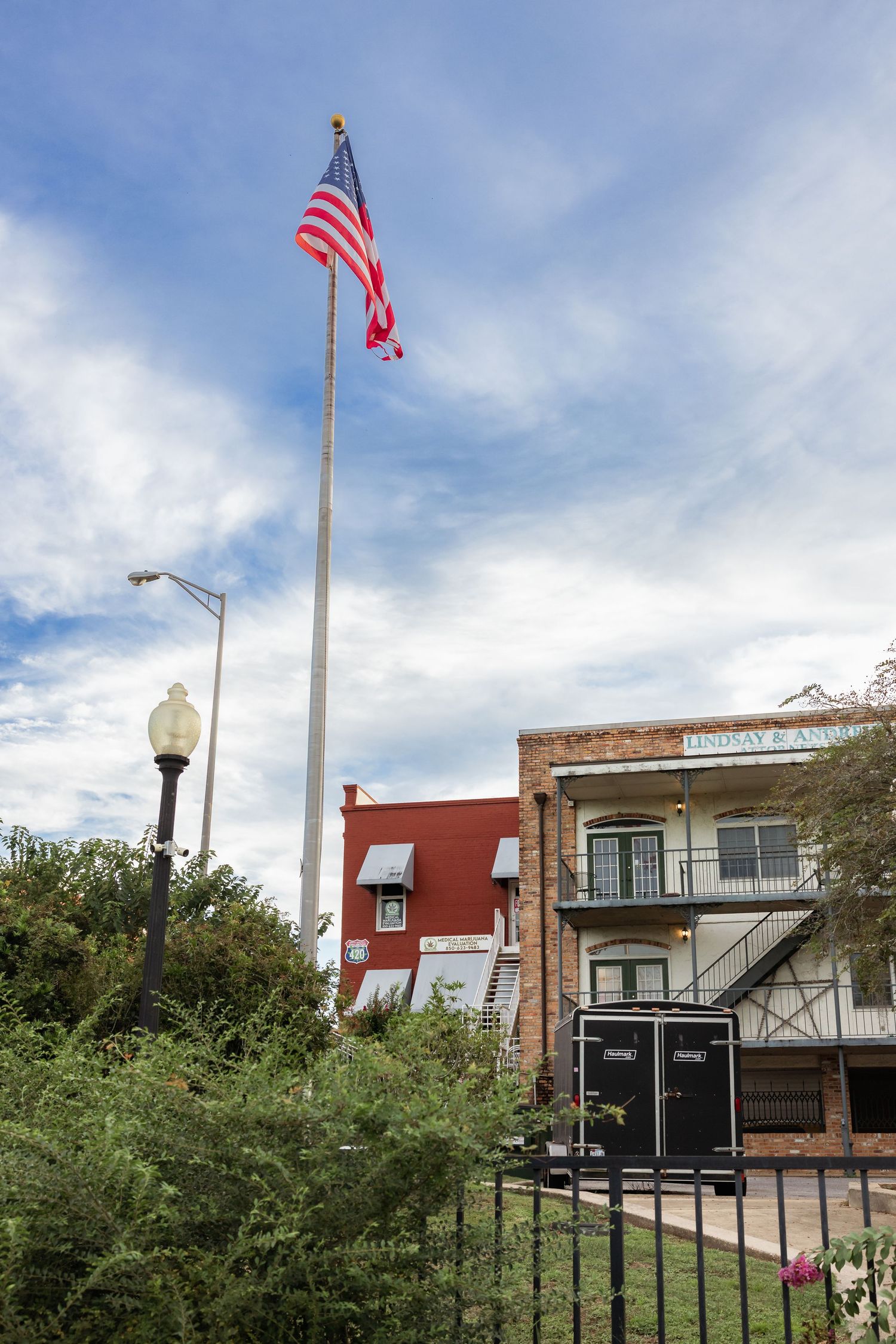 American flag on tall flagpole beside brick buildings and street lamp under partly cloudy sky in small town USA.
