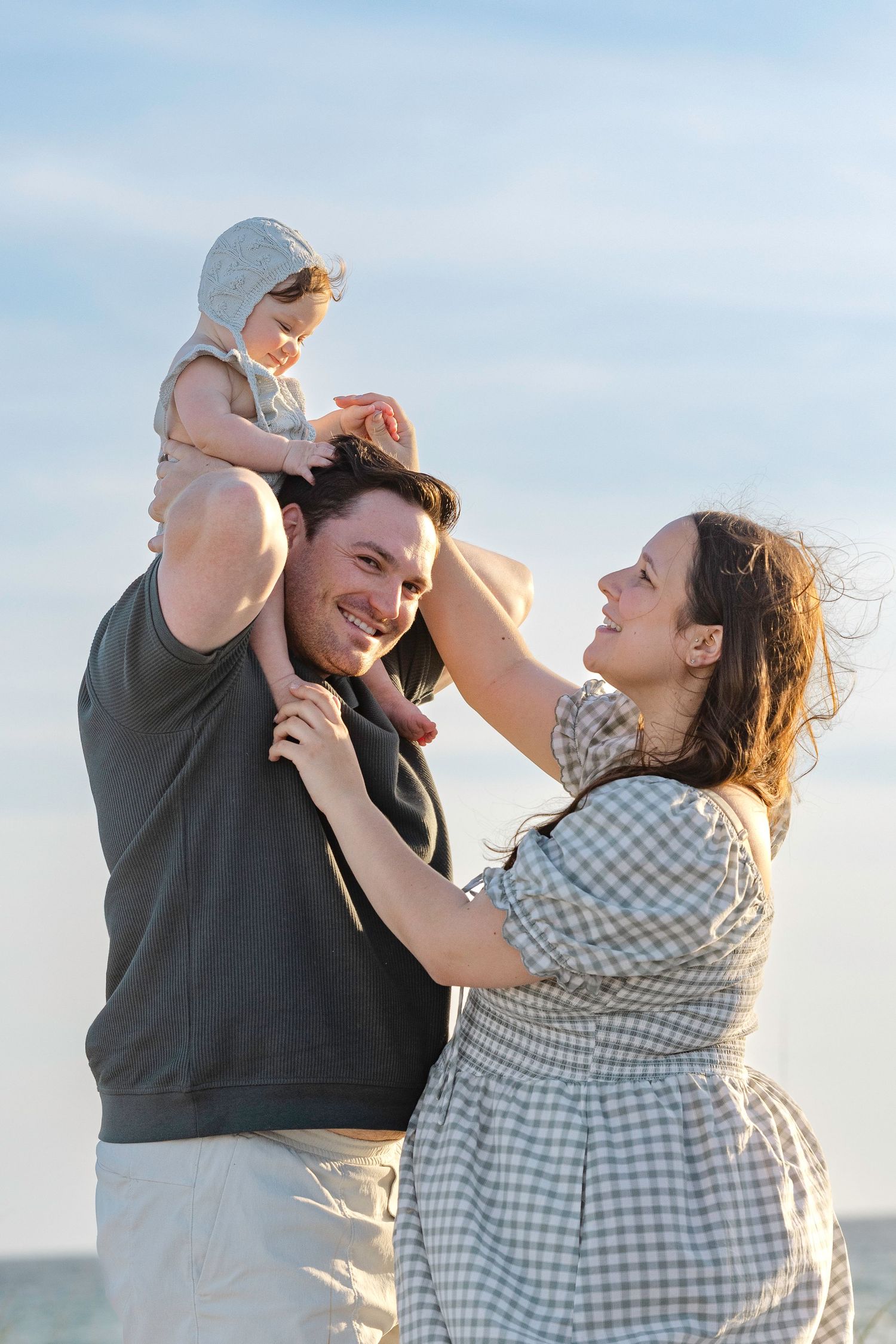 Happy family with father holding baby on shoulders while mother smiles outdoors under blue sky.