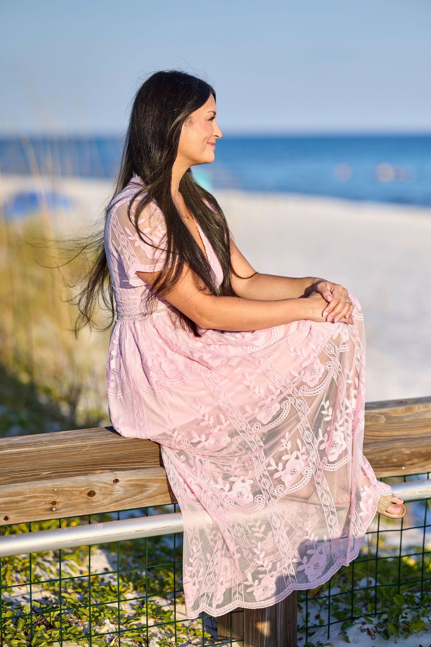 Woman in pink lace maxi dress sitting on wooden fence at sunny beach with ocean views.