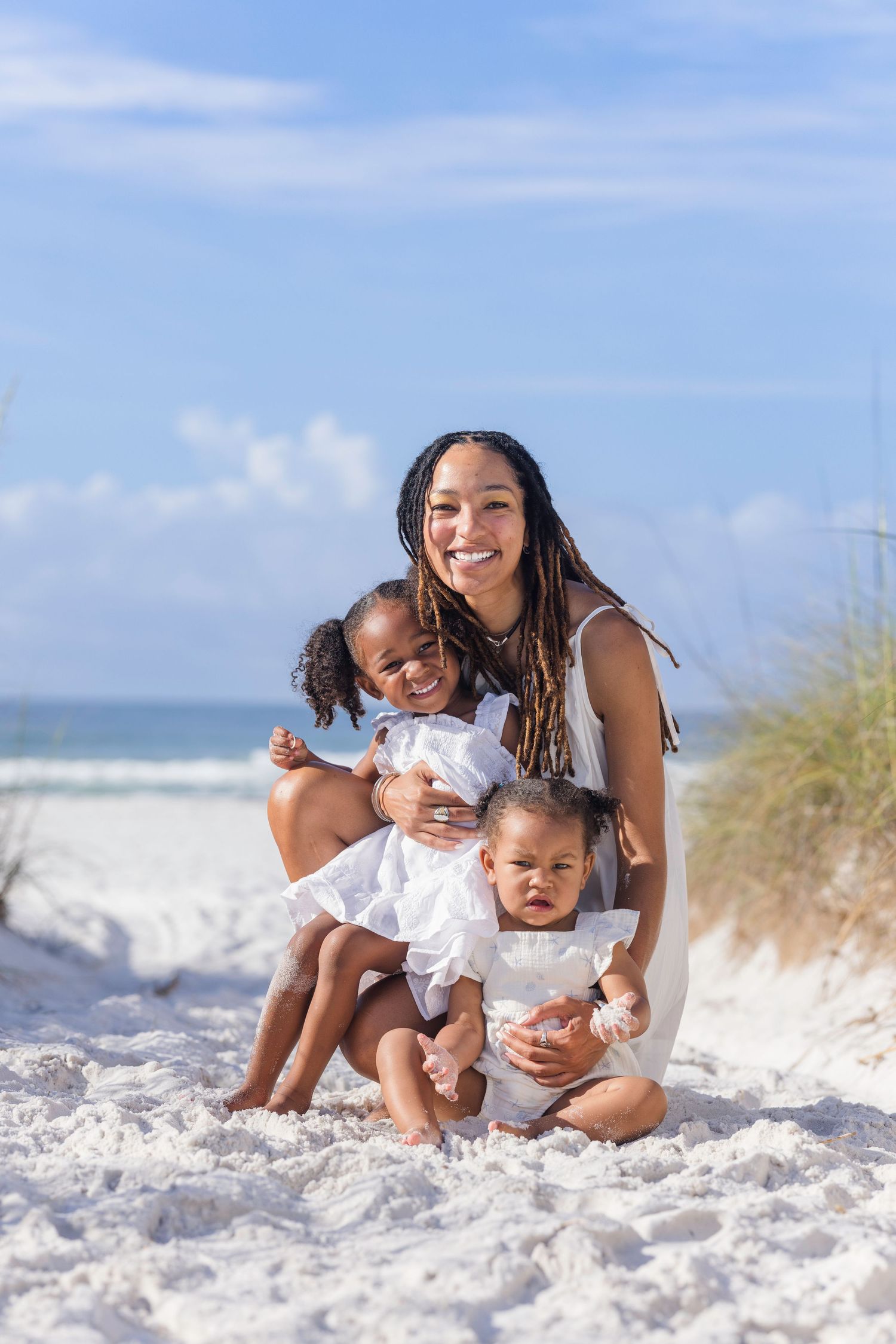 A smiling mother with braids sits on white sand beach holding two young daughters in white dresses.