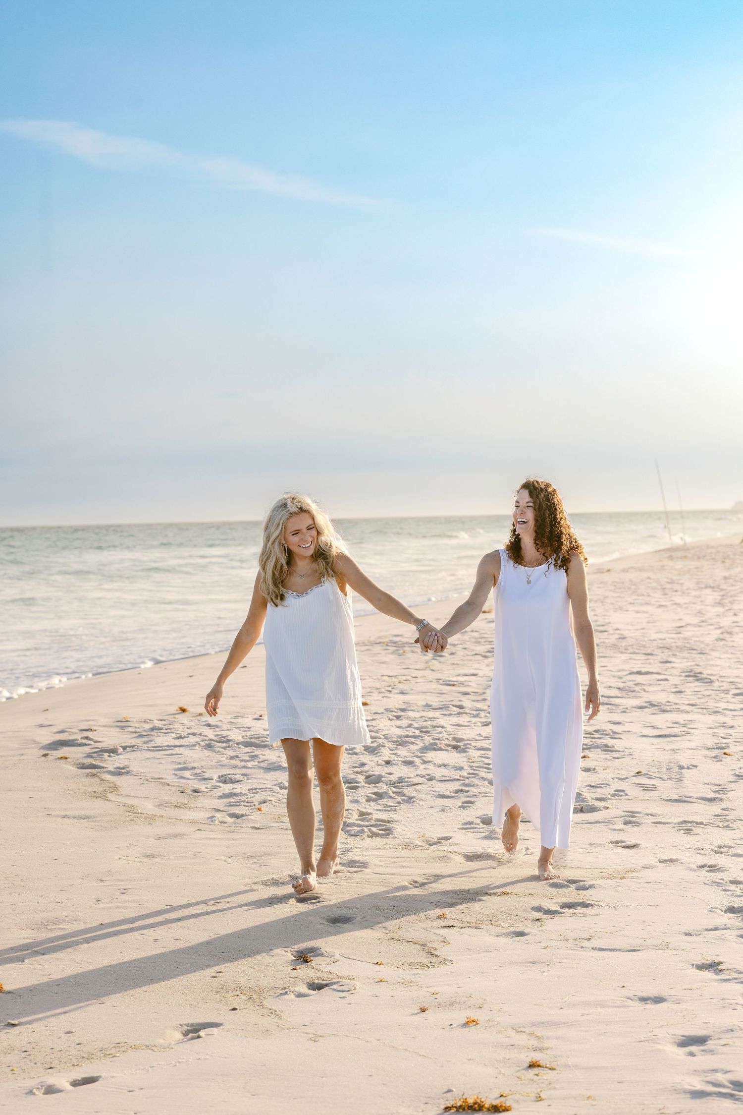 Two women in white dresses hold hands walking on a sunny beach with calm ocean waves in the background.