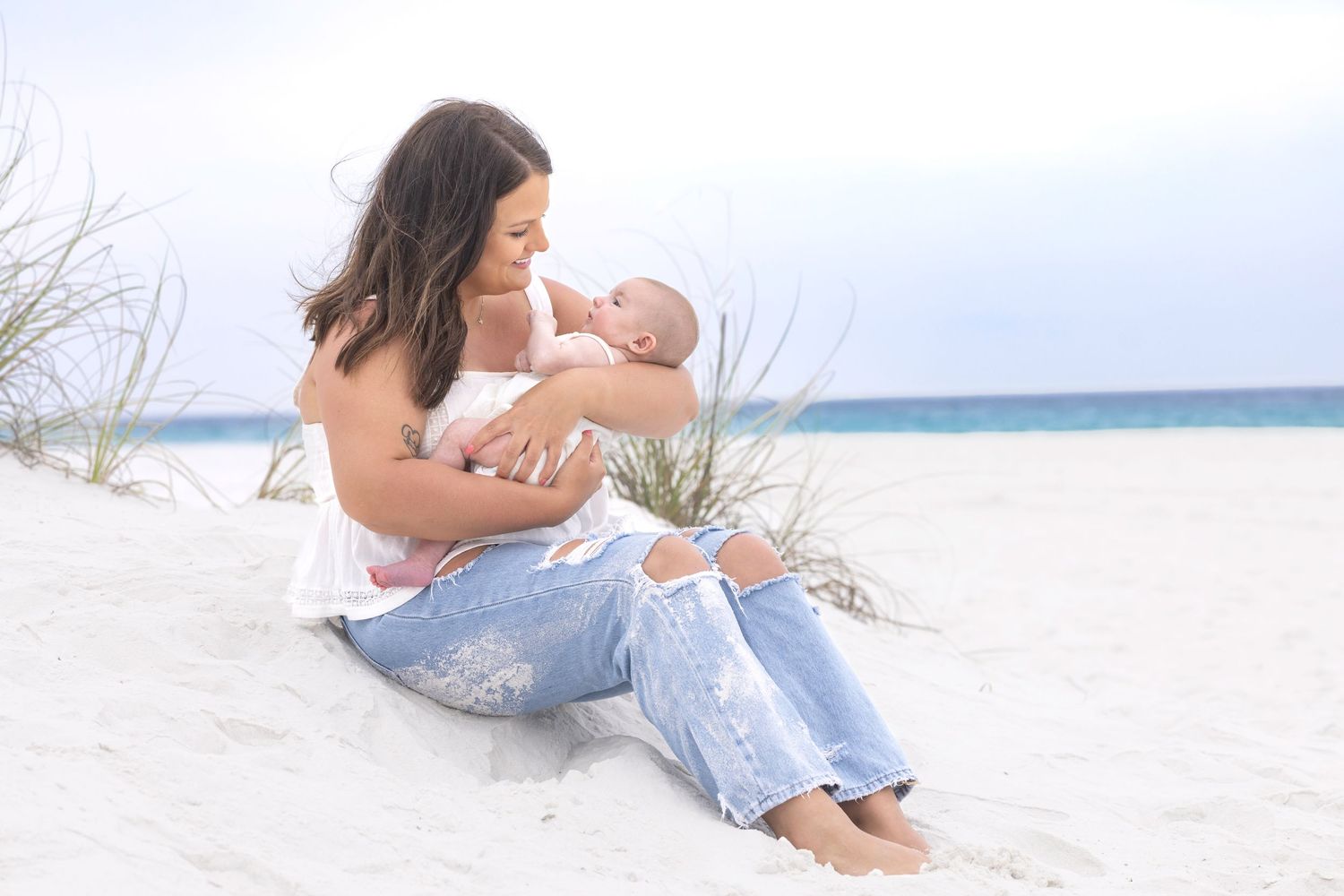 Mother breastfeeding newborn baby on white sand beach, sitting on dunes in ripped jeans and white top.