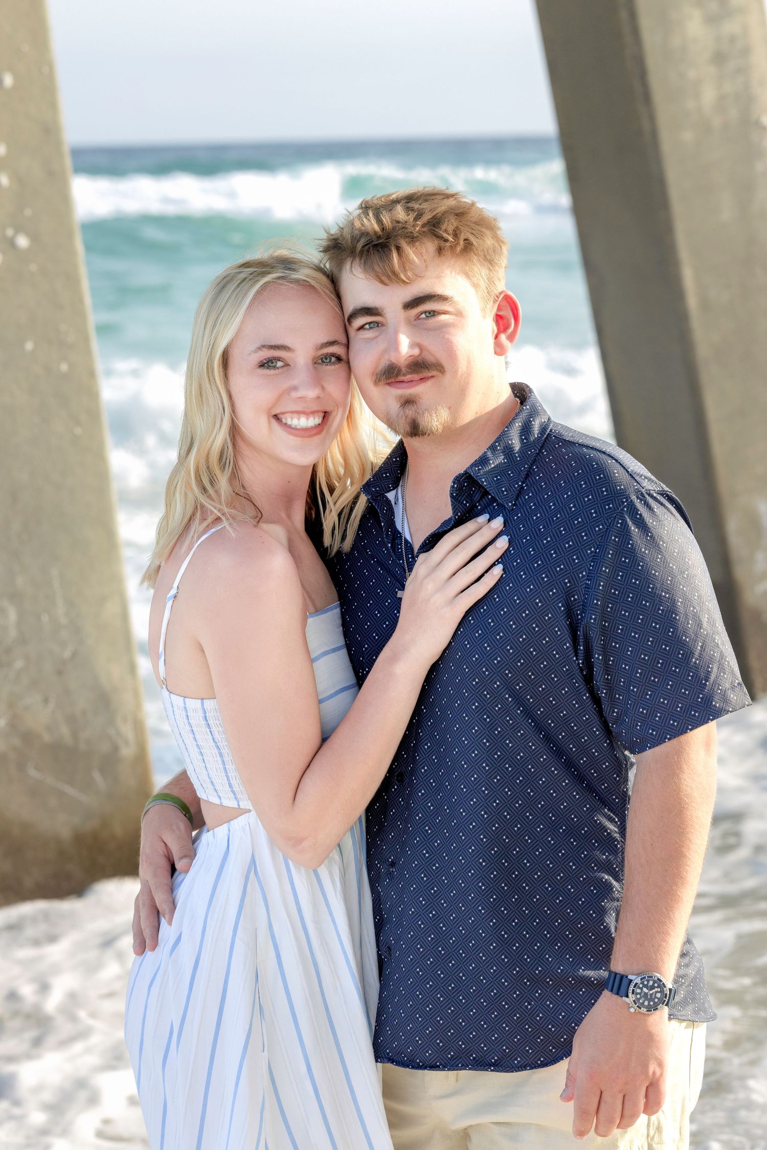 Smiling young couple posing together under a beach pier with ocean waves in the background.