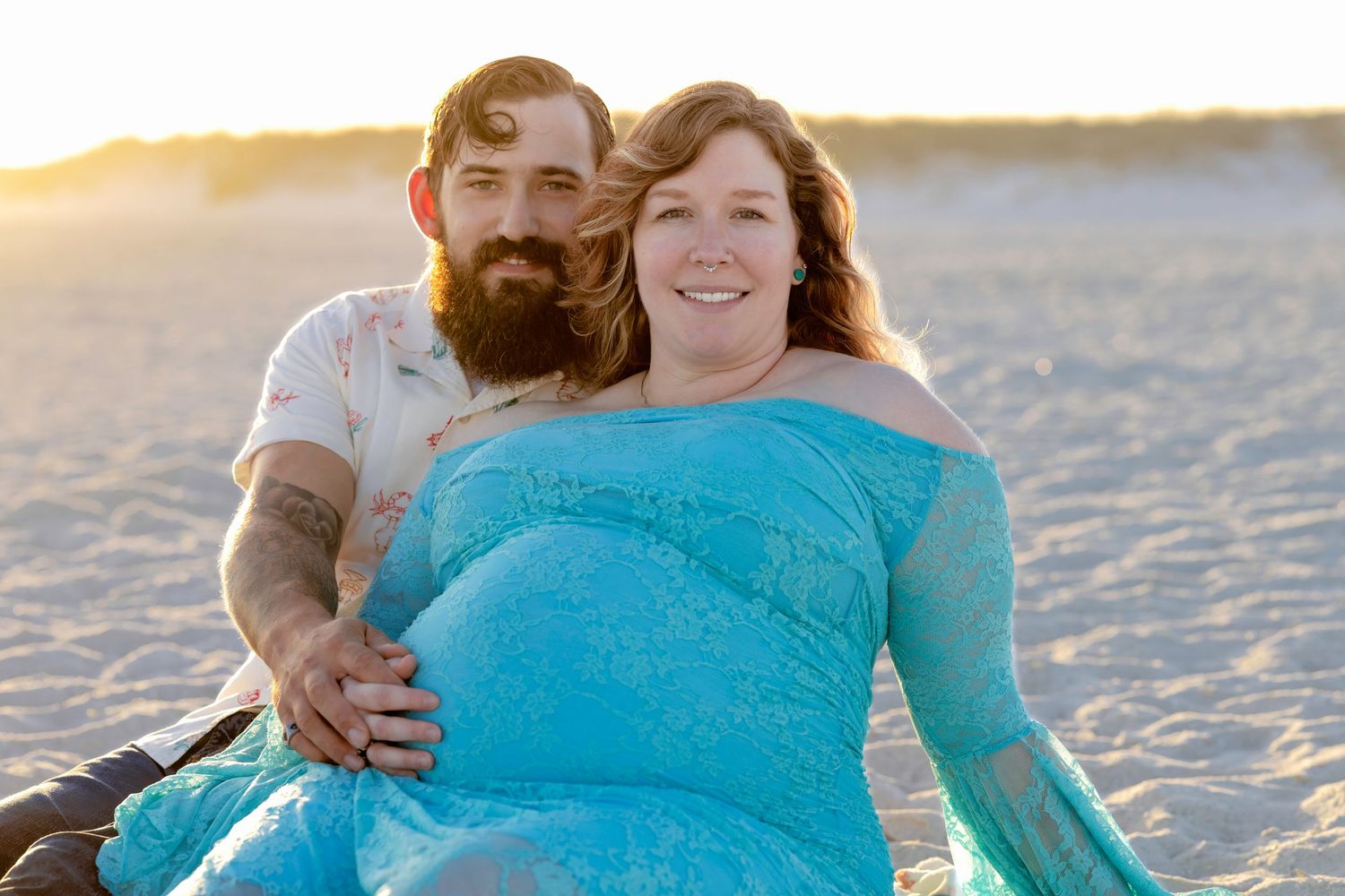 Pregnant couple smiling together on beach at golden hour, woman in turquoise maternity gown.