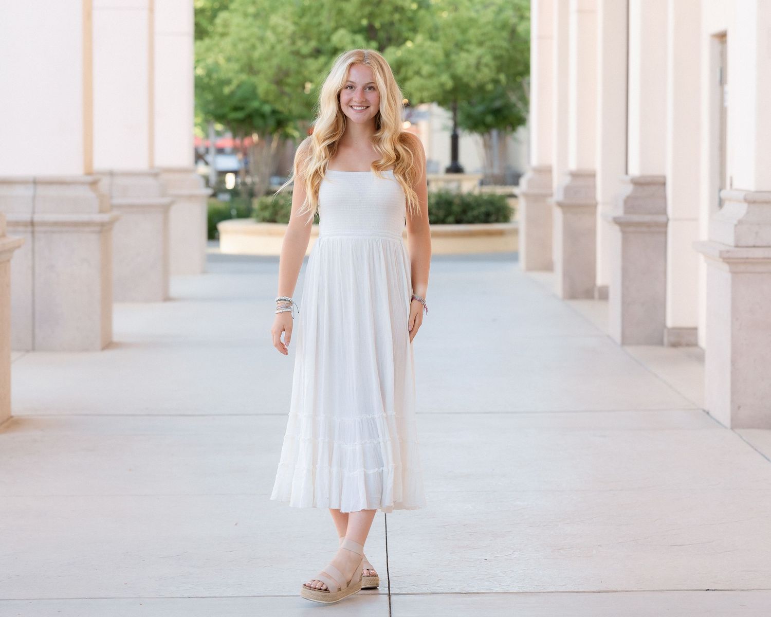 Young blonde woman posing in a white maxi dress between stone columns.