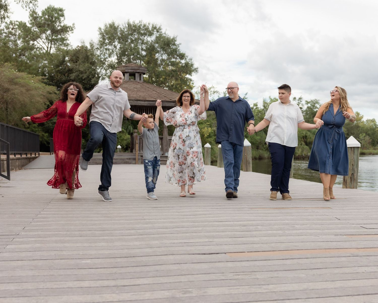 Family of seven holding hands and walking on a wooden dock near a lake, smiling and enjoying a sunny day outdoors.