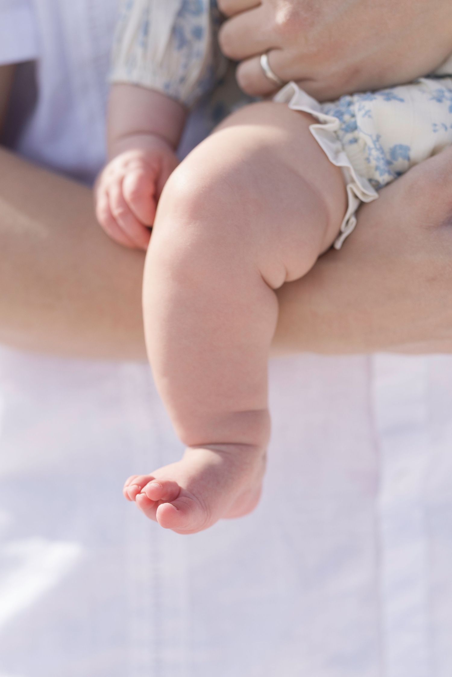 Close-up of a cute baby's chubby leg and tiny toes being held by a parent wearing a white outfit.