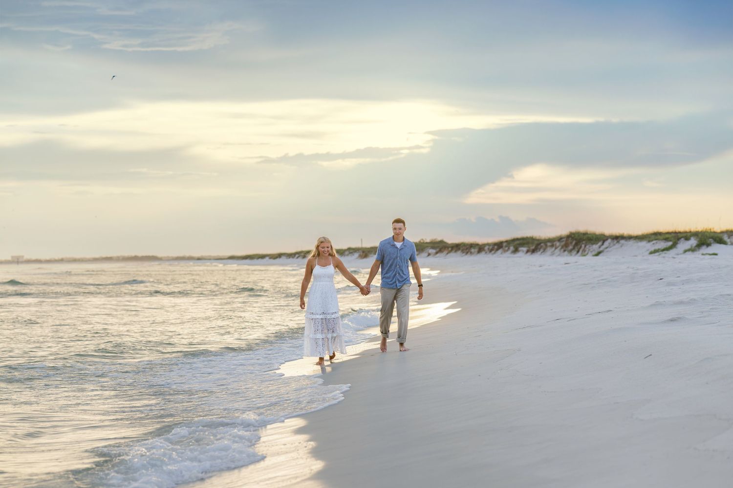 Couple holding hands walking along a sunny beach shoreline at sunset in white and blue outfits.