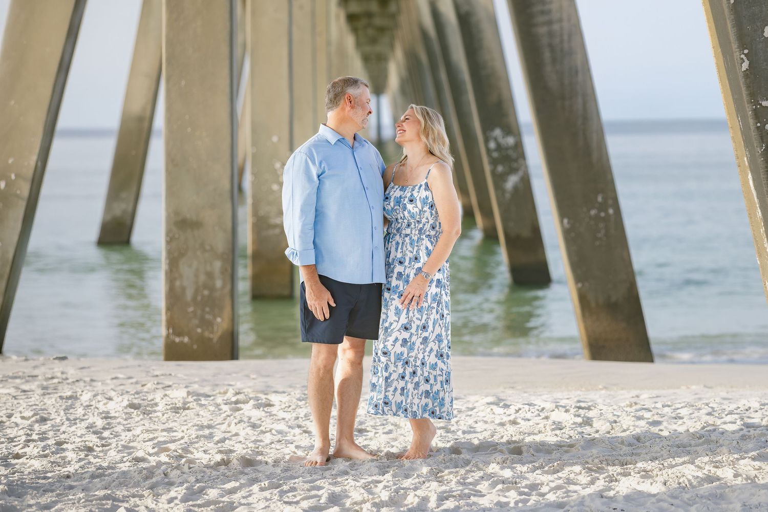 Couple standing together on a sandy beach beneath a wooden pier, man in blue shirt and woman in floral dress.