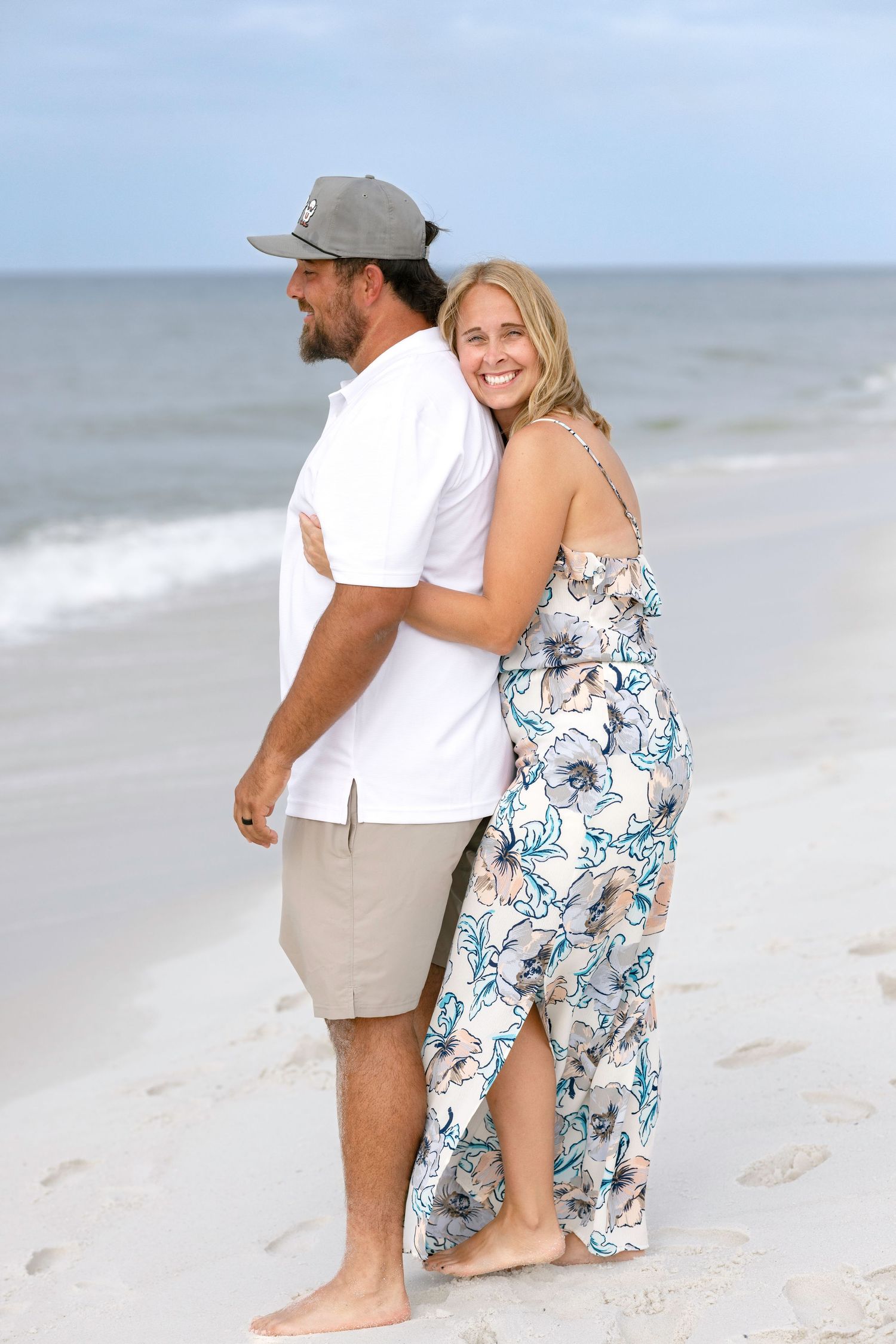 Smiling couple hugging on sandy beach shoreline, woman in floral jumpsuit, man in white shirt and cap.