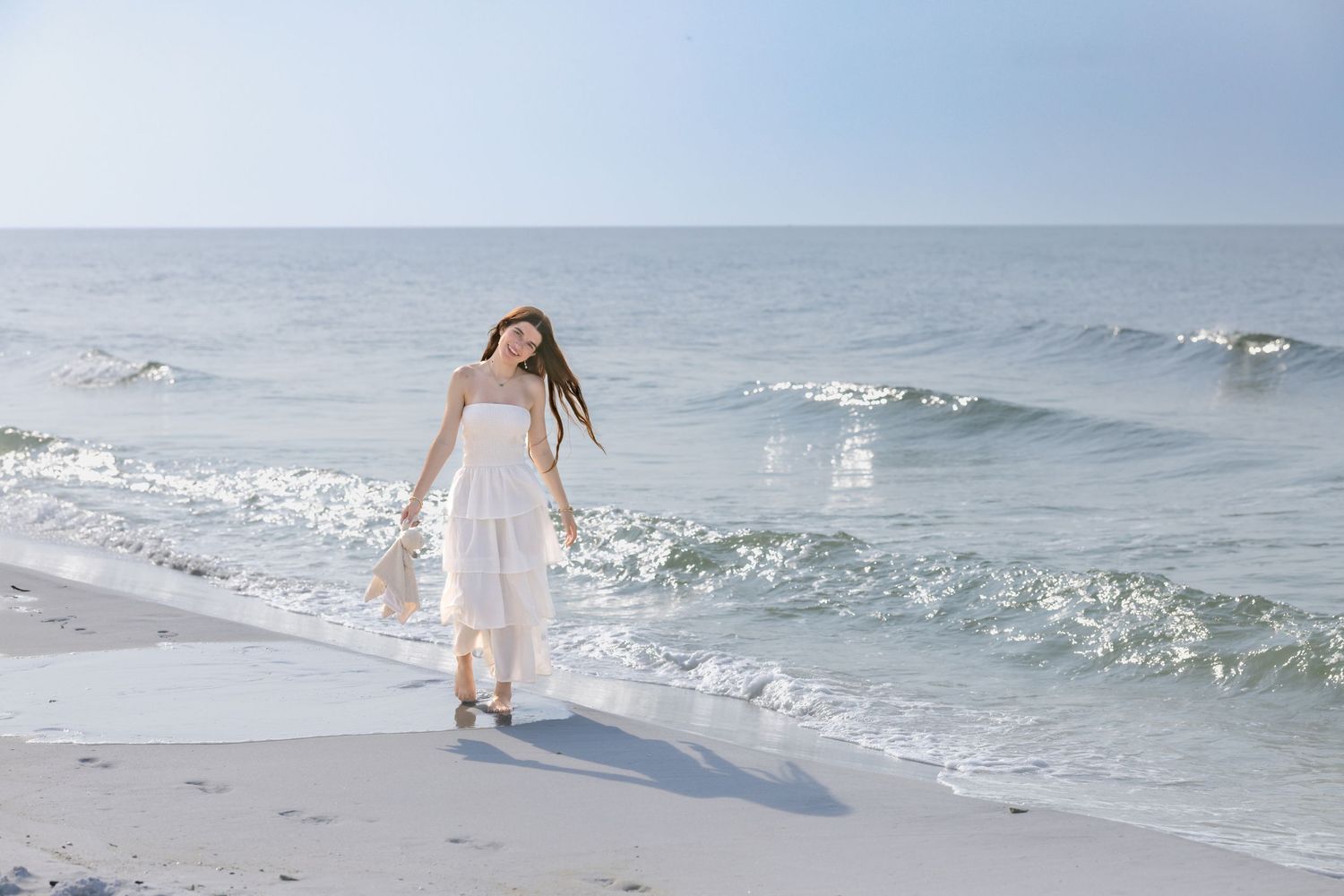 A young woman in a white sundress walks along the shoreline with ocean waves gently rolling behind her.