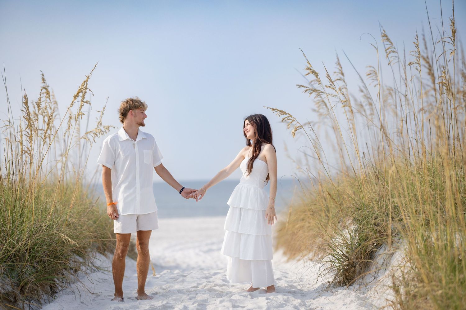Couple in white outfits holding hands on a sandy beach path surrounded by sea oats under a clear blue sky.