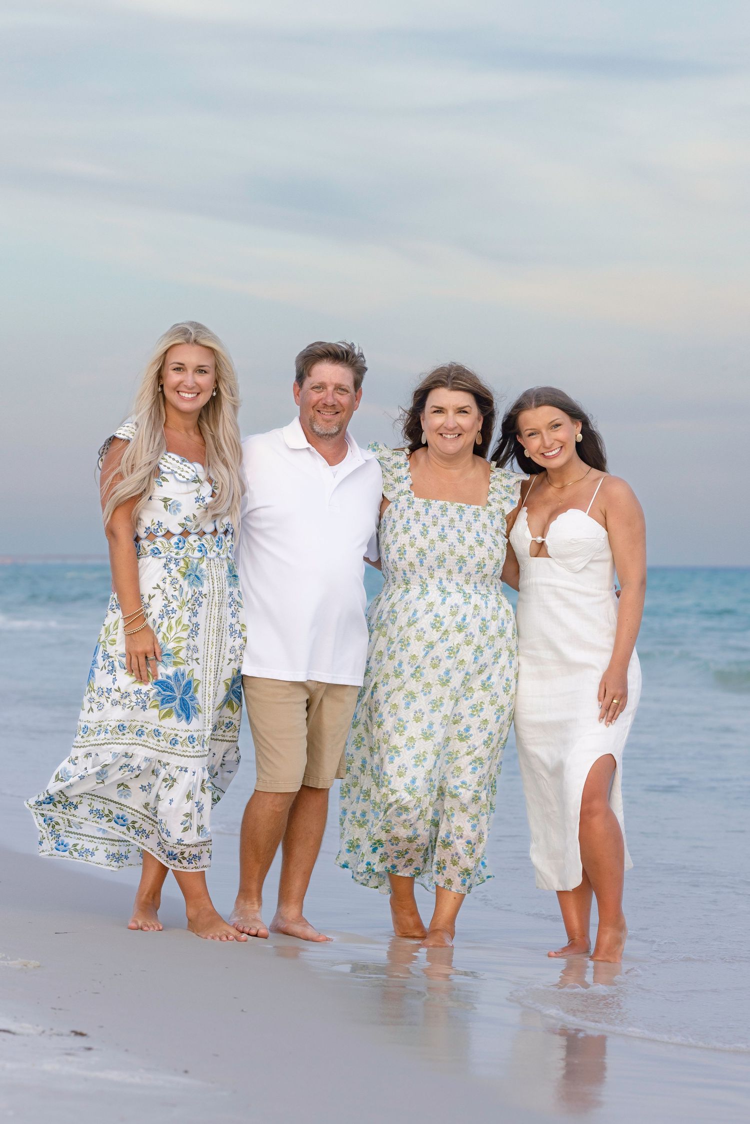 Family of four posing together on a sandy beach at sunset, wearing coordinated outfits in blue and white tones.