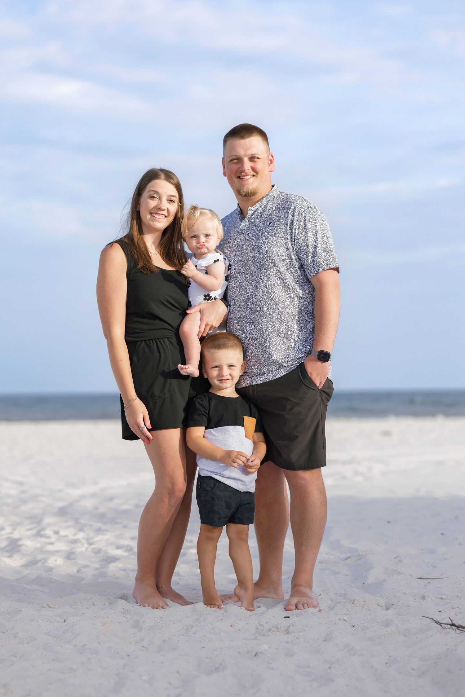 Family of four posing on white sand beach, parents holding toddler with young boy standing in front.