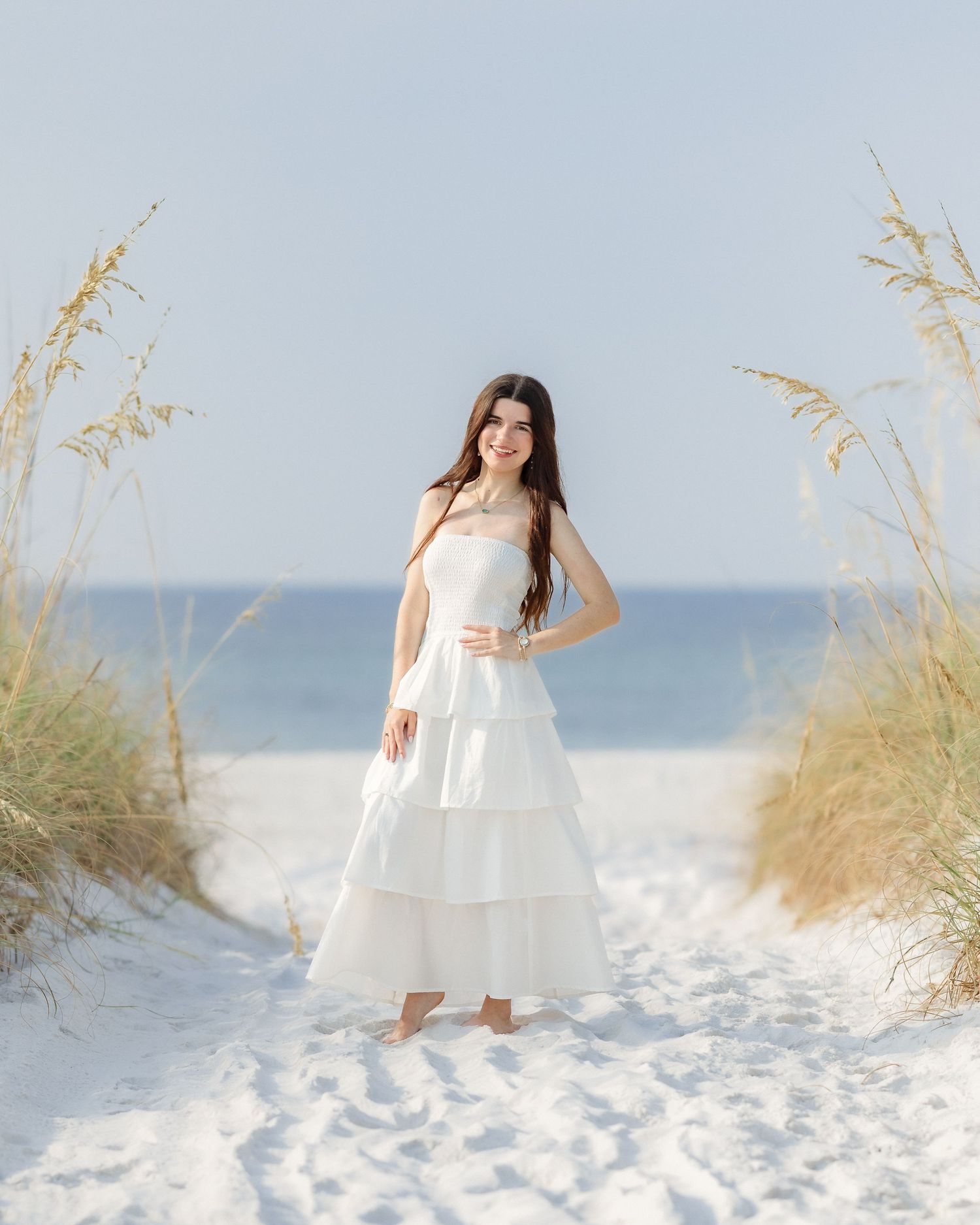 Young woman in white tiered dress stands barefoot on white sand beach path framed by sea oats.