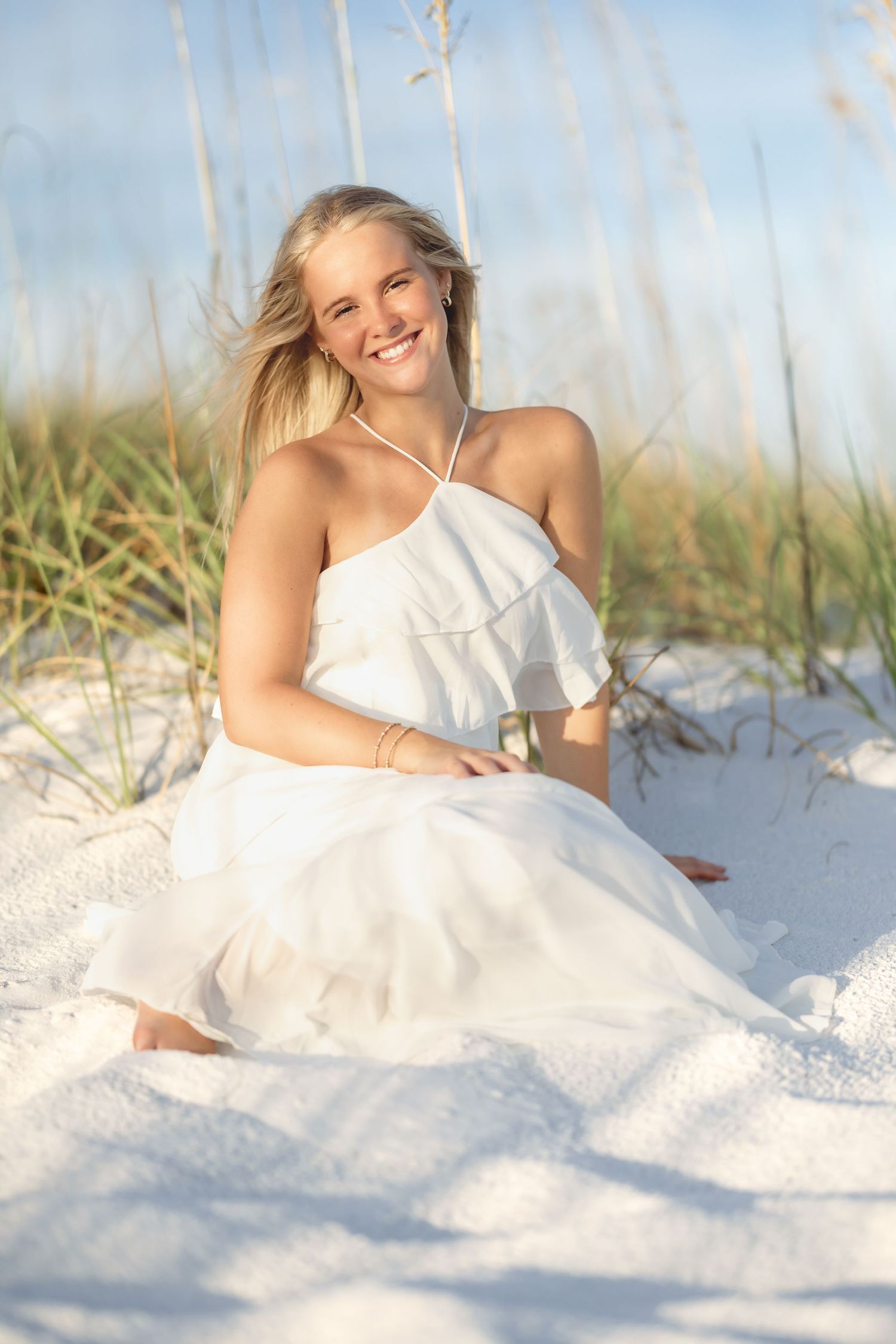 Smiling blonde woman in white halter dress sitting on white sand beach dunes with sea oats in background.