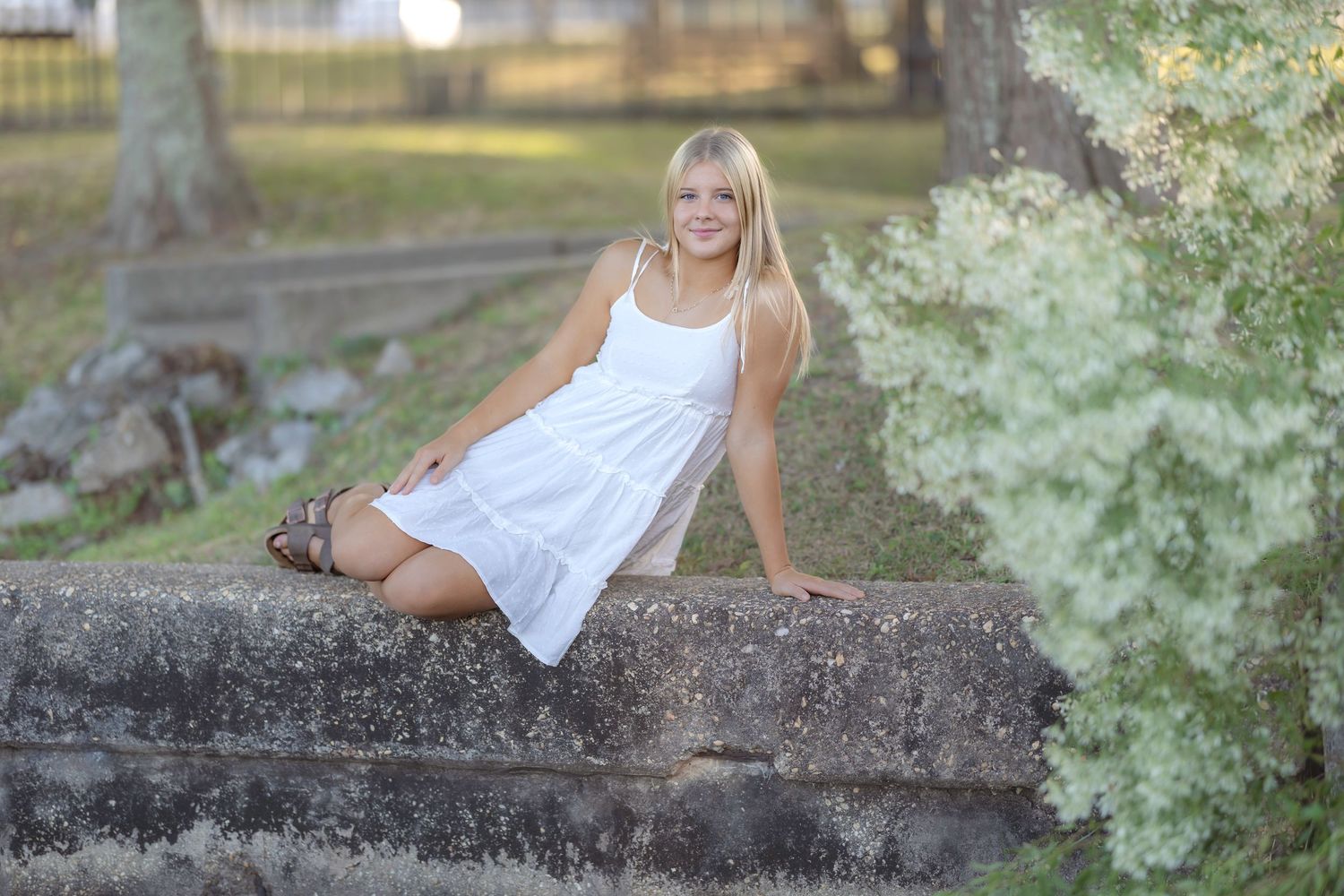 Young blonde woman in white sundress sitting on stone steps outdoors surrounded by lush green trees and foliage.