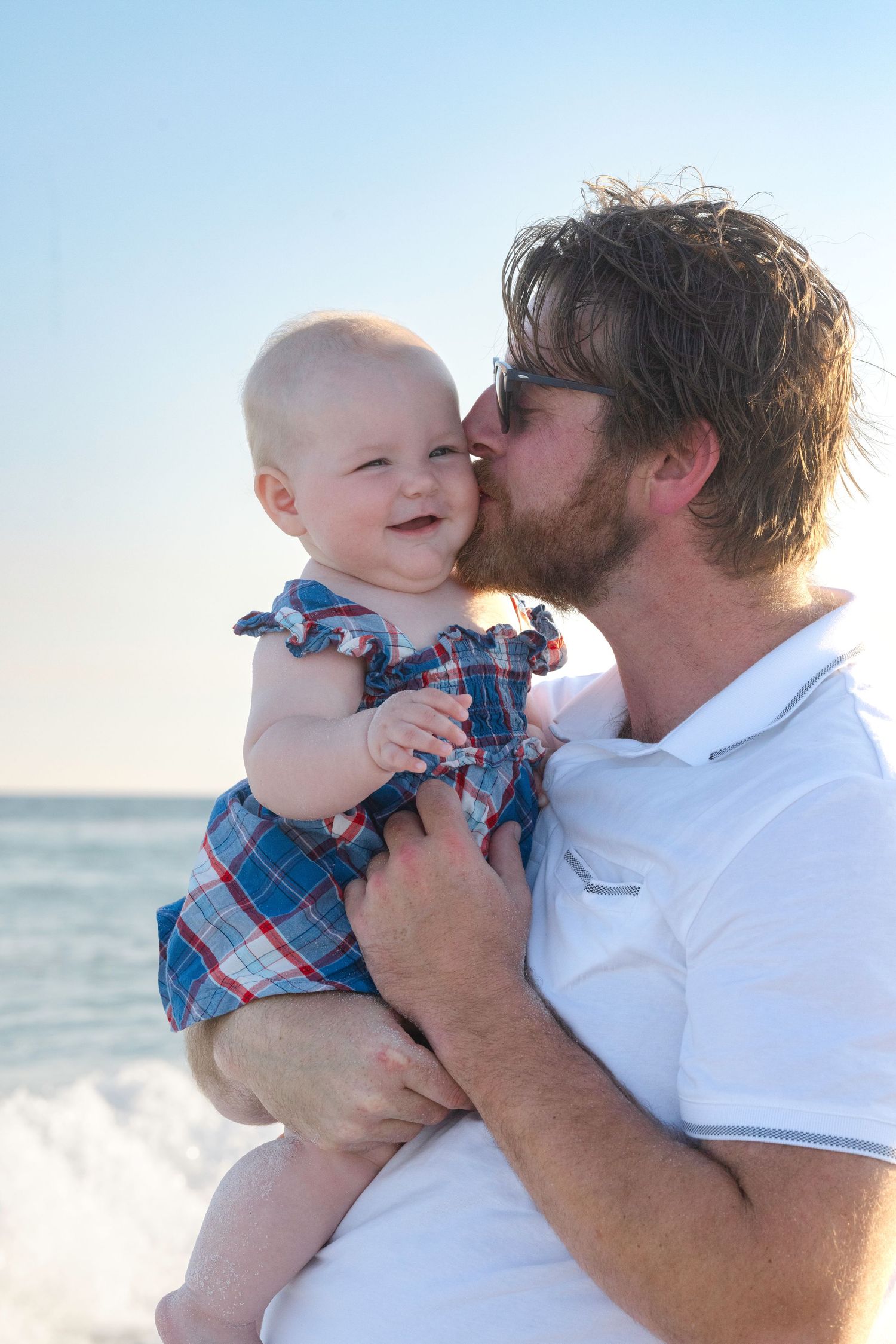 A bearded father kisses his smiling baby girl on the cheek while holding her at the beach.