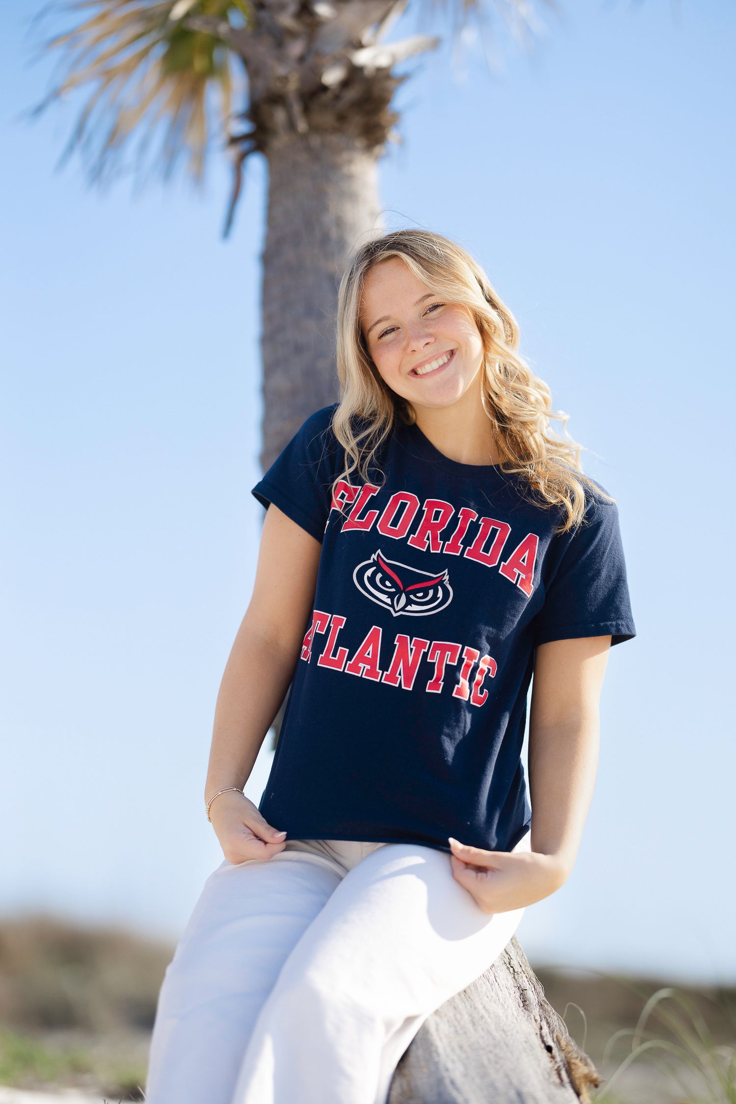 Person wearing Florida Atlantic navy t-shirt and white pants poses outdoors with palm trees in background.