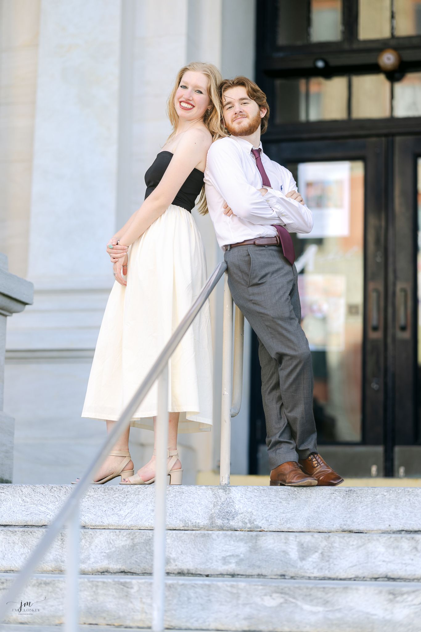 A couple poses playfully on white marble steps in formal attire.